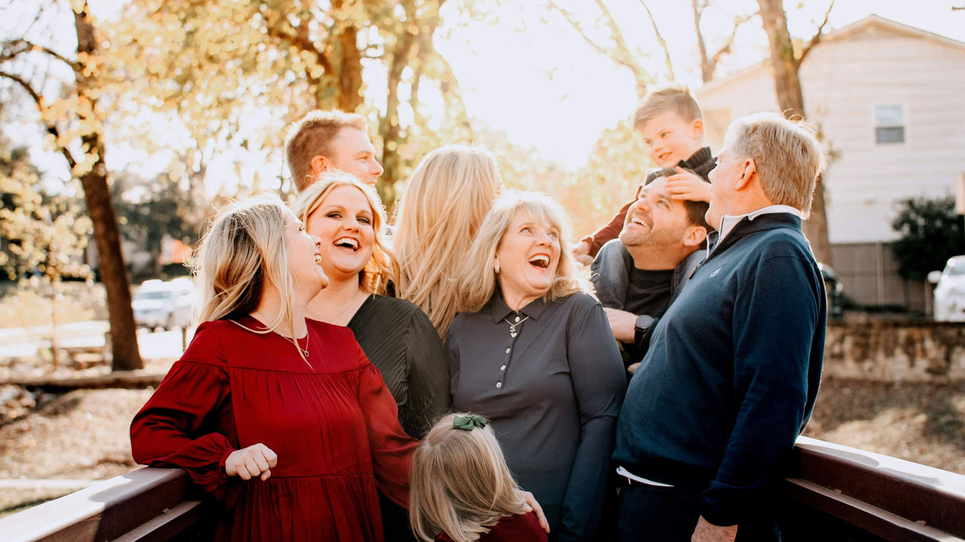 Större familj samlas tätt tillsammans på en träbro utomhus i varmt solljus, med flera generationer som står nära varandra i en avslappnad miljö.