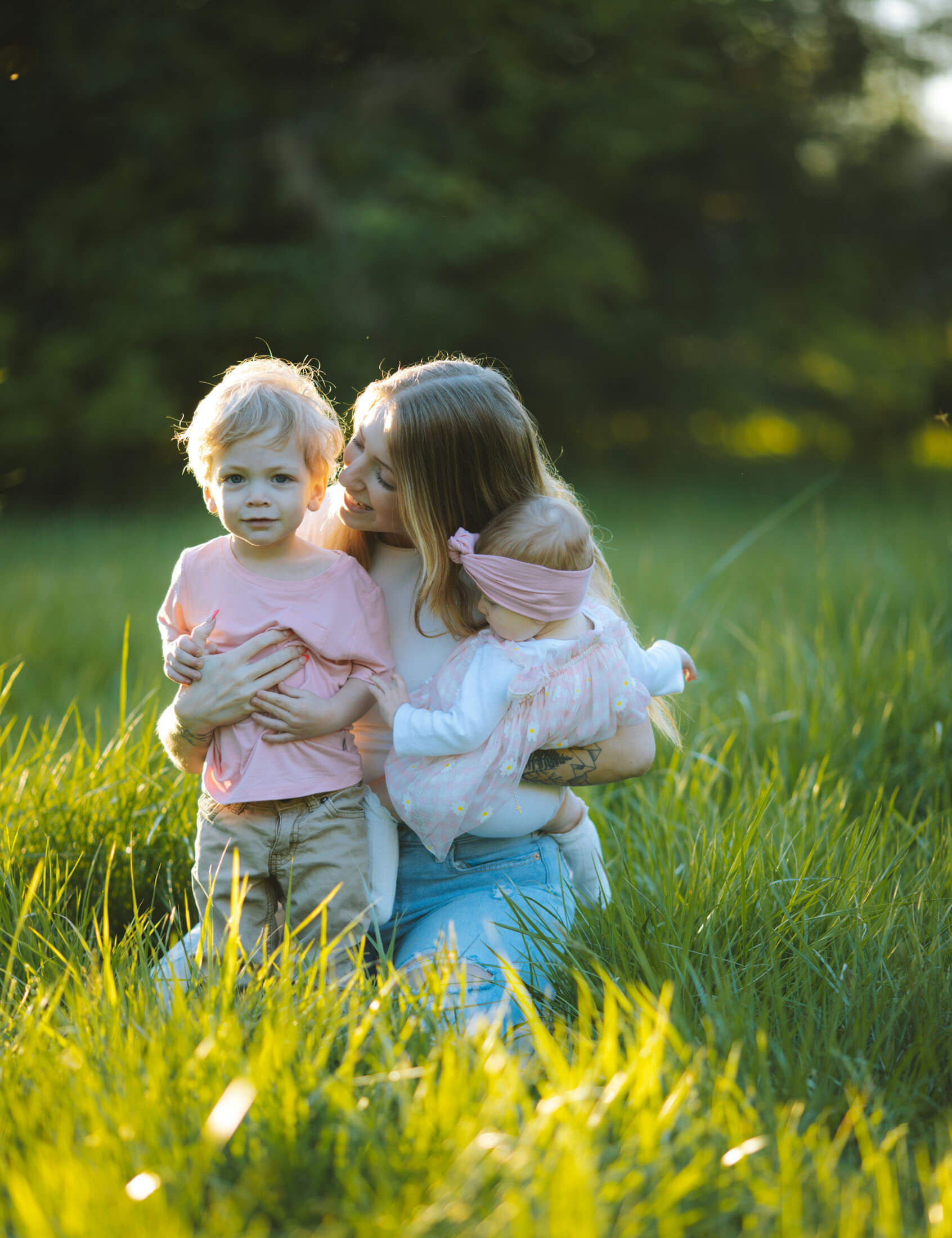 Mamma sitter i högt gräs och håller två små barn i famnen, fotograferad i mjukt kvällsljus med suddig grön bakgrund.