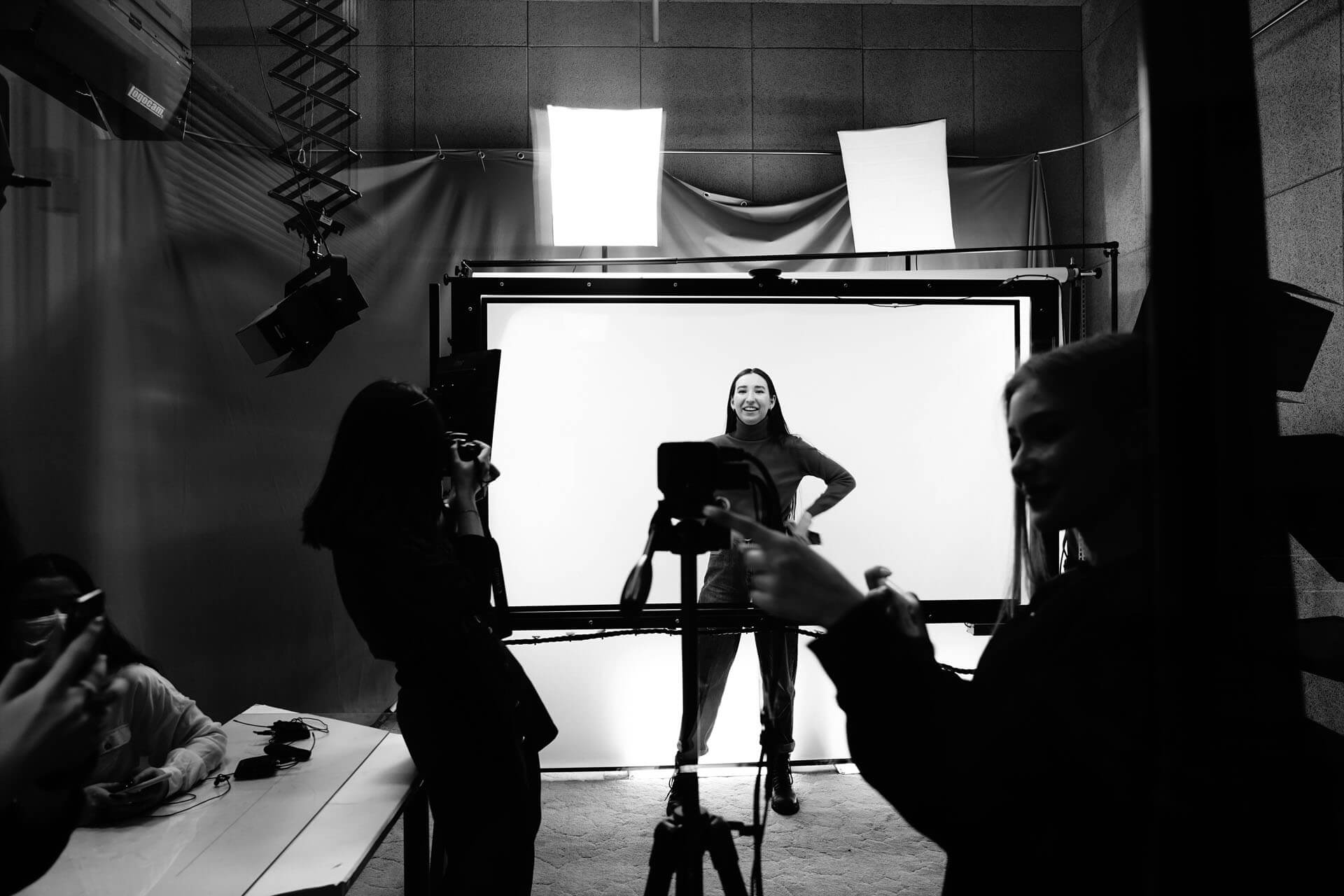 Fotostudio i svartvitt där flera personer står runt en upplyst bakgrund medan en person poserar framför ljuskällor och kameror i en kreativ inspelningsmiljö.