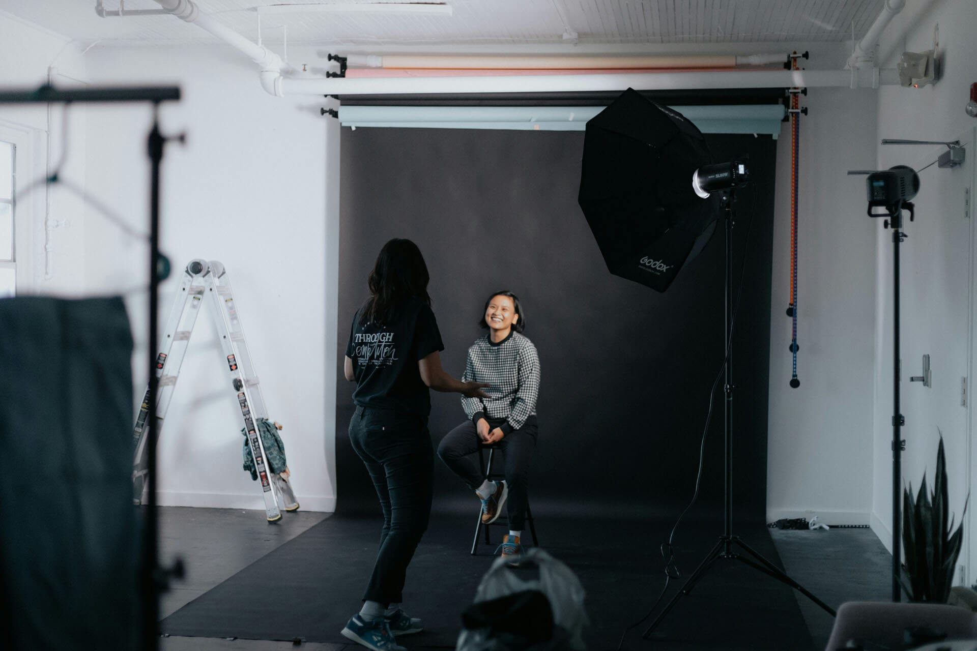 Fotostudio med svart bakgrund där en person sitter på en pall och fotograferas, omgiven av studiobelysning, stativ och utrustning i en arbetsmiljö.