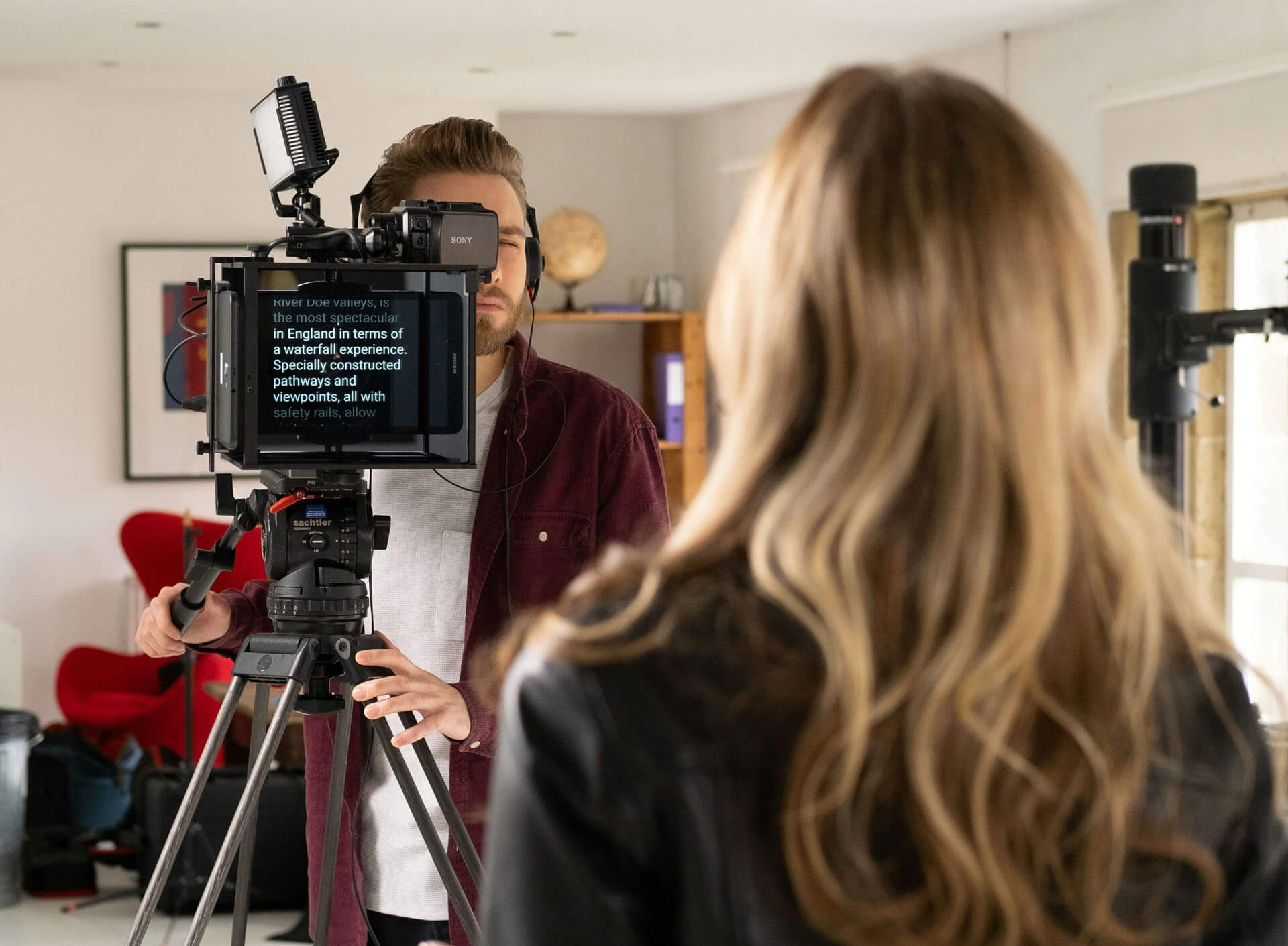 Person står vid kamera och teleprompter under videoinspelning i studio, med mikrofon, stativ och kontorsliknande miljö i bakgrunden.