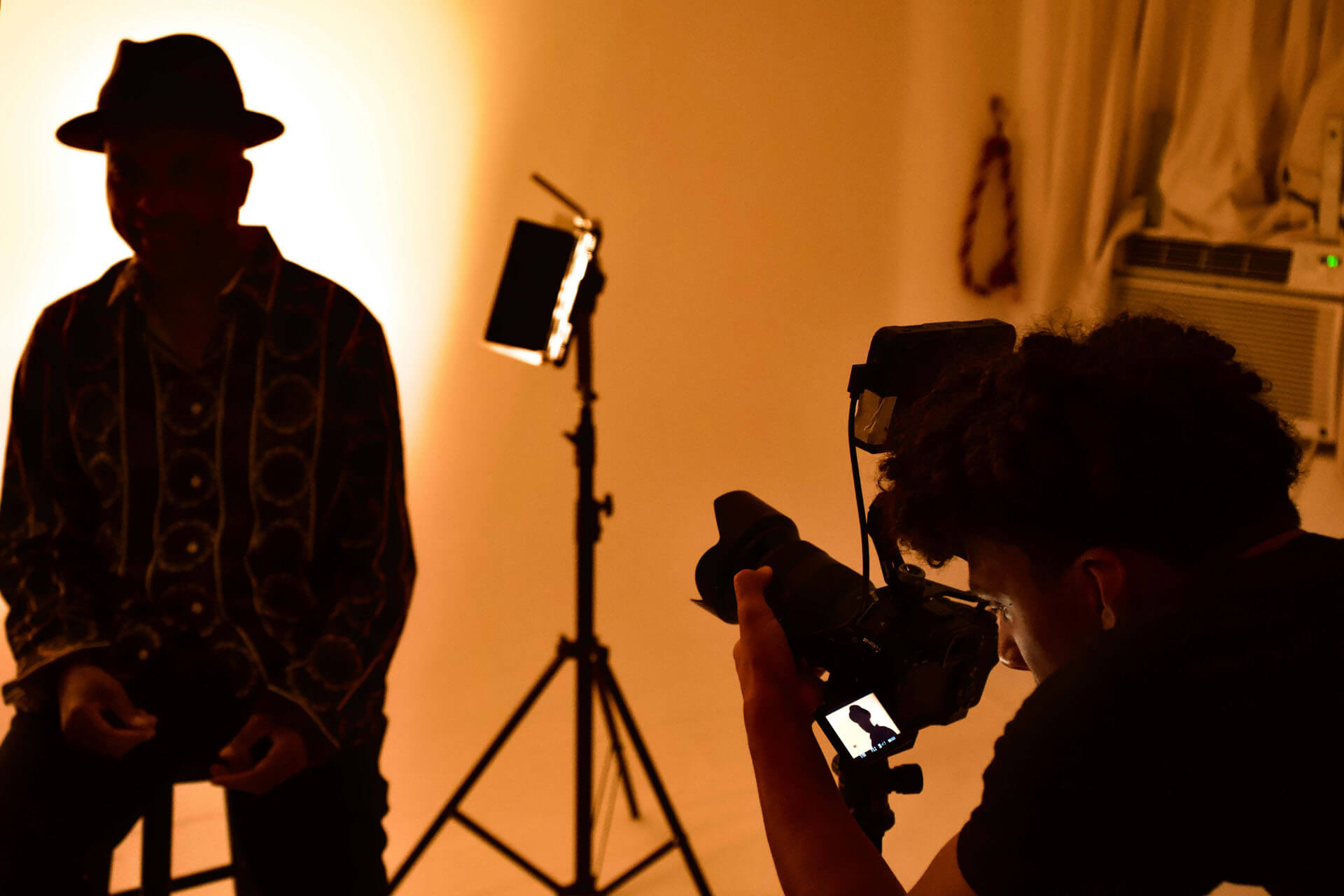 Fotograf arbetar i studio och riktar kamera mot person som sitter på stol framför starkt varm bakgrundsljus, med ljusstativ och utrustning synliga.