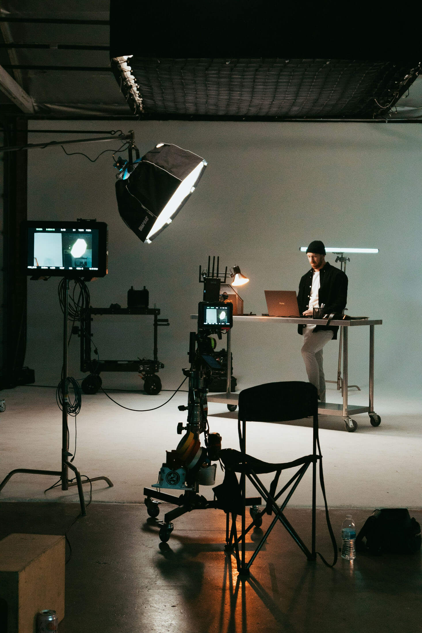 Studiomiljö med kameror, ljusstativ och monitorer där en person står bakom ett bord och arbetar vid laptop, medan utrustning och stolar syns i förgrunden.