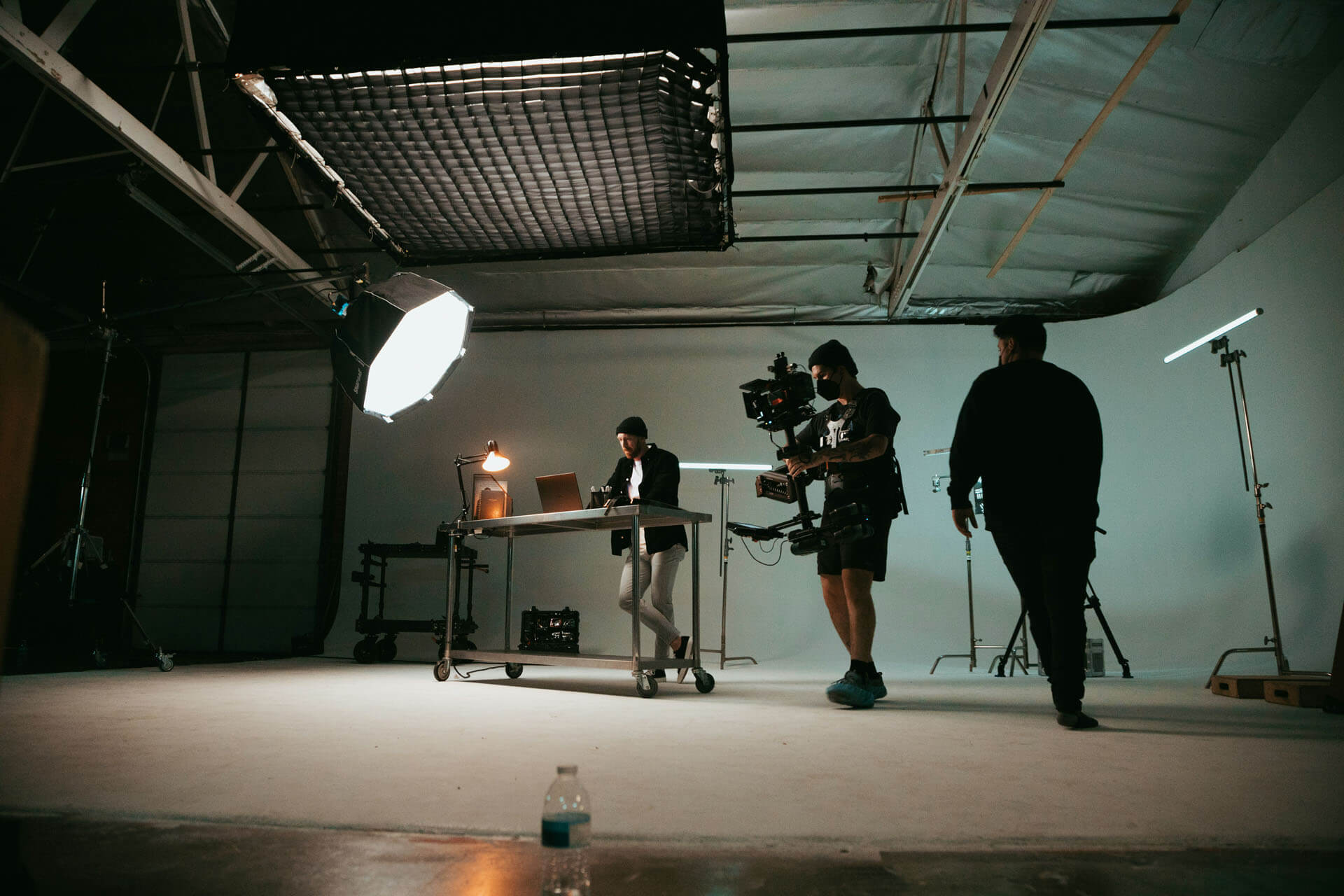 Filmteam arbetar i en studio där en person sitter vid ett bord med laptop medan en kamerafotograf med rigg filmar, omgiven av stora ljuskällor och utrustning.