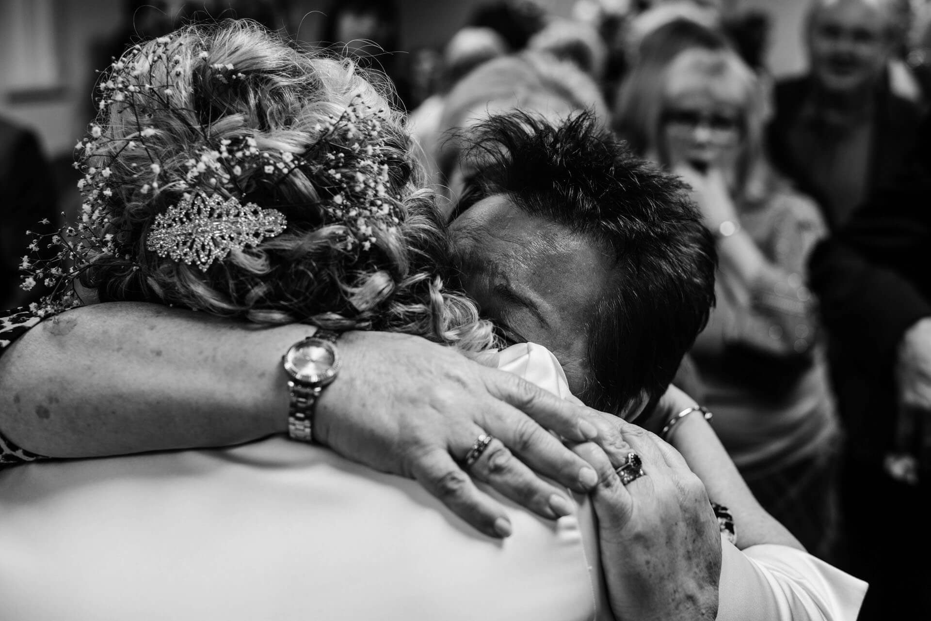 Svartvitt foto där två personer omfamnar varandra nära, med fokus på händer, hårdetaljer och smycken i en dokumentär och intim stund omgiven av suddig publik i bakgrunden.