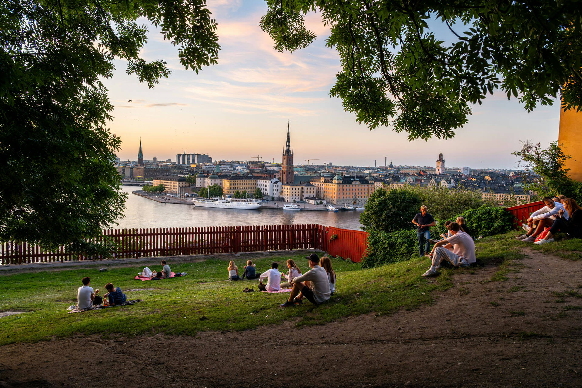 Utsikt över Stockholm i kvällsljus från en grön park där flera personer sitter på gräset och ser ut över vatten, stadssiluett och kyrktorn omgivna av träd.