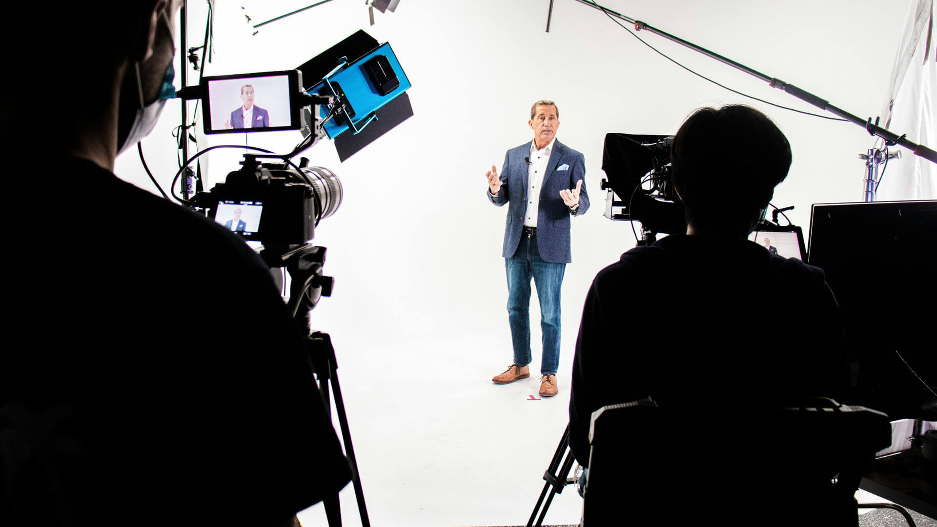 Studioinspelning där en person står framför vit bakgrund och talar mot kamera, omgiven av belysning, kamerariggar och teknisk utrustning i en professionell inspelningsmiljö.