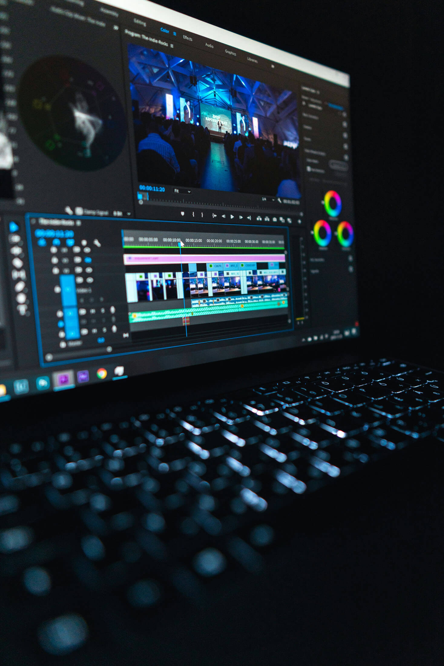 Datorskärm visar videoredigeringsprogram med tidslinje, färgpaneler och klipp från ett event, fotograferad i mörk arbetsmiljö.