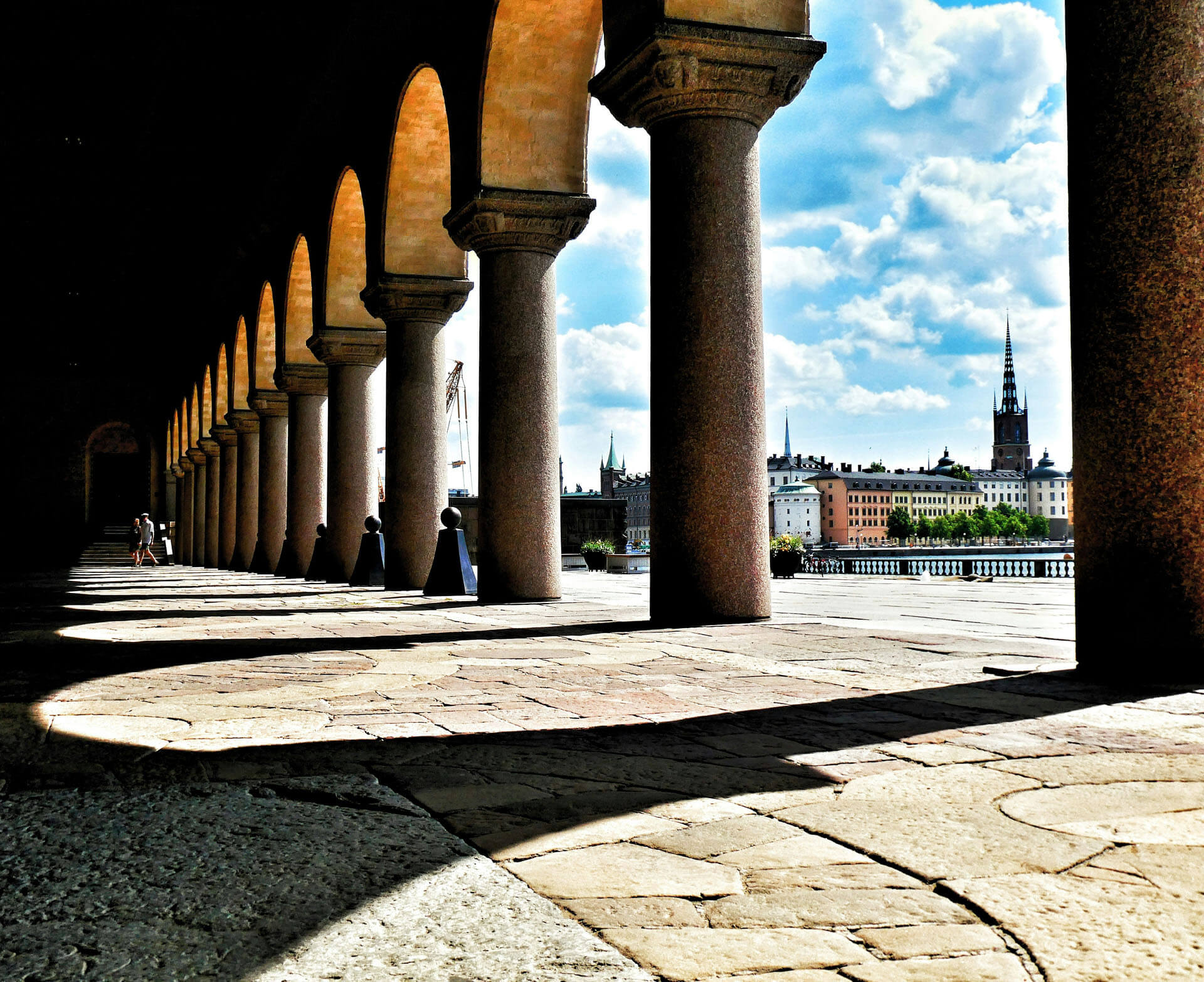 Rad av höga stenpelare och valv i skugga leder ut mot en solbelyst utsikt över Stockholm, med kyrktorn och byggnader synliga i bakgrunden.
