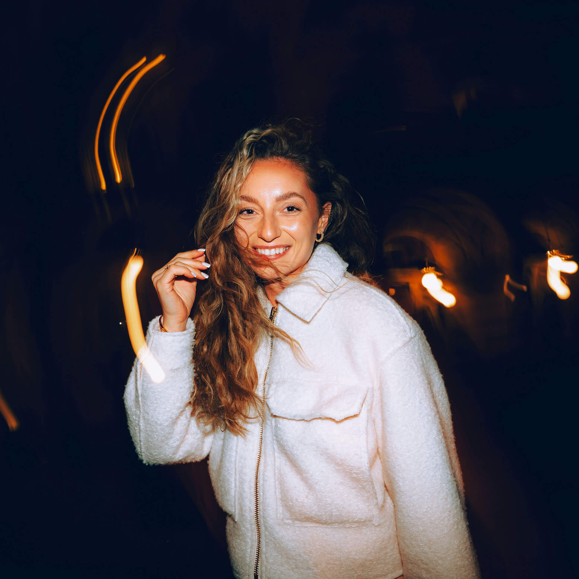 TFP-fotografering utomhus i Stockholm där en kvinna med långt brunt hår och ljus jacka ler mot kameran i kvällsmiljö. Med ena handen i håret och varma ljusspår i bakgrunden skapas ett kreativt modellfoto som kombinerar mode, rörelse och personlighet.
