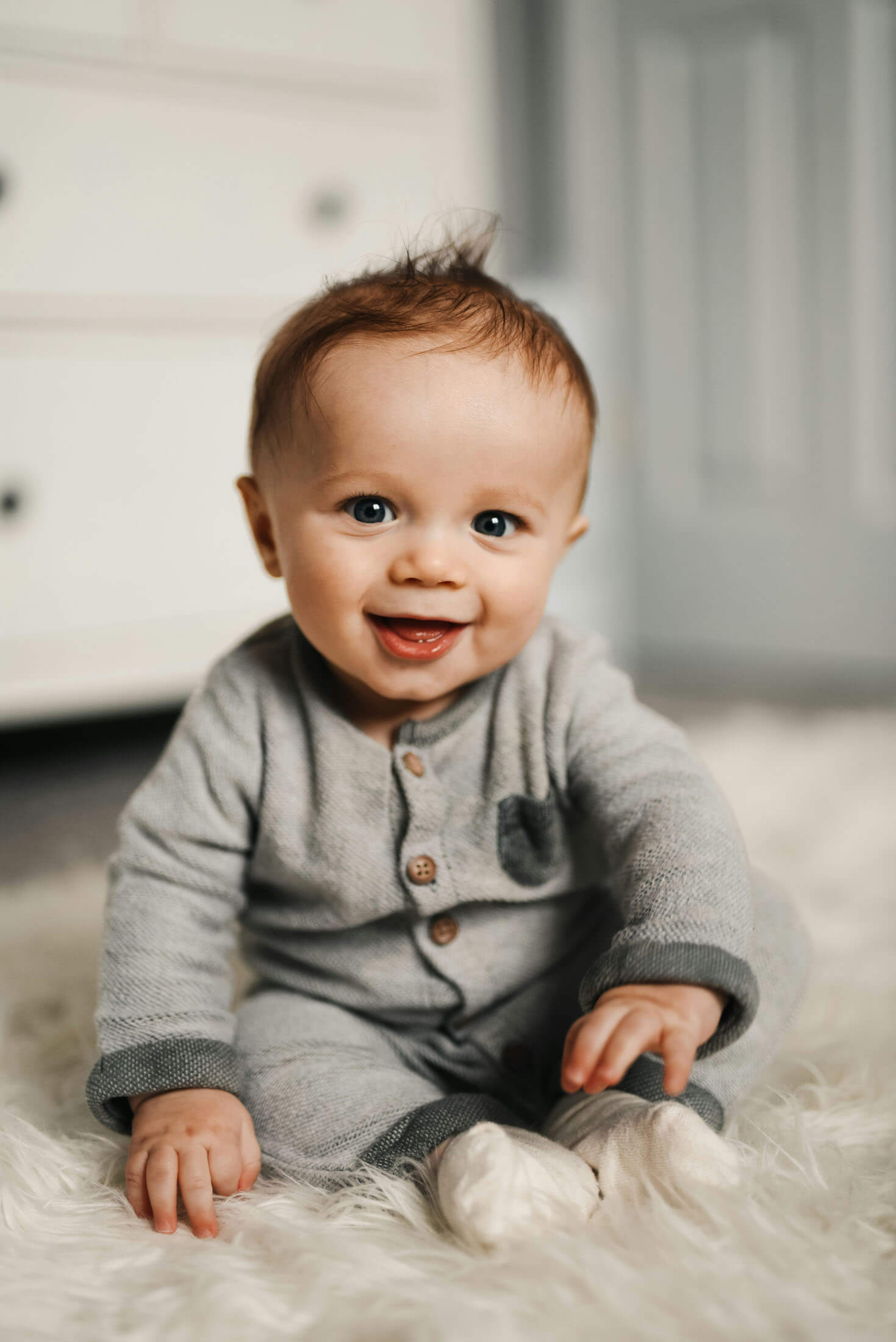 Ett spädbarn sitter på golvet och ler mot kameran. Barnet bär en grå långärmad outfit och har händerna framför sig för stöd. Bakgrunden är mjukt suddig och ger bilden en varm och hemtrevlig känsla.