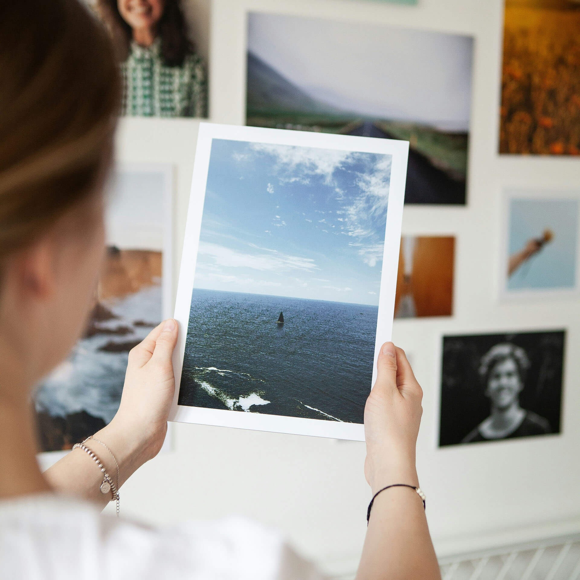  Person håller upp ett utskrivet fotografi av hav och himmel framför en vägg med flera inramade bilder i bakgrunden.