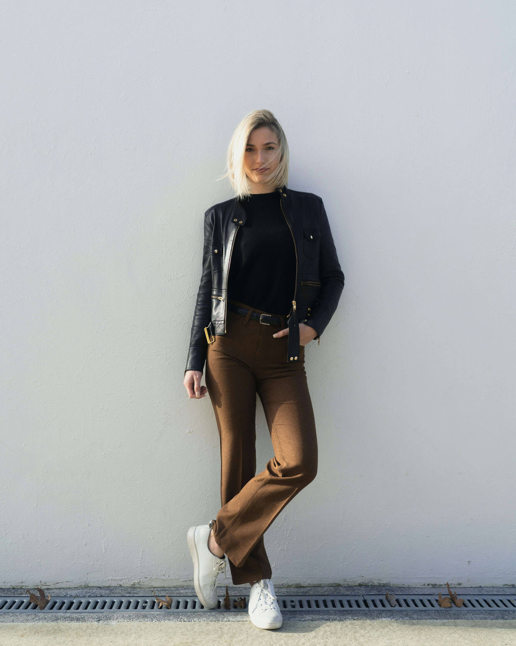 En person står lutad mot en ljus vägg med händerna i fickorna. Personen bär en mörk långärmad tröja, bruna byxor och ljusa skor. Bilden har en minimalistisk stil med rena linjer och gott om tom yta runt motivet.