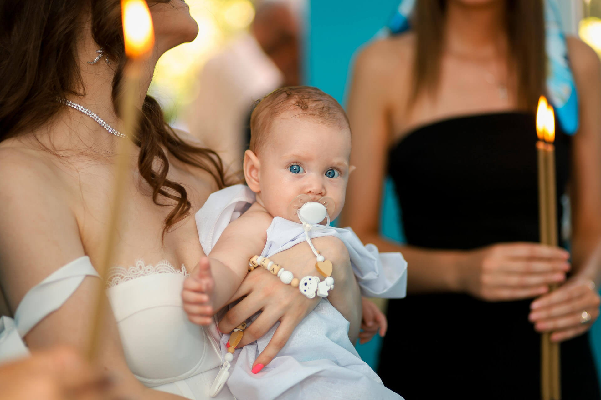 Bebis med napp i famnen under dopceremoni med tända ljus som hålls upp i bakgrunden.