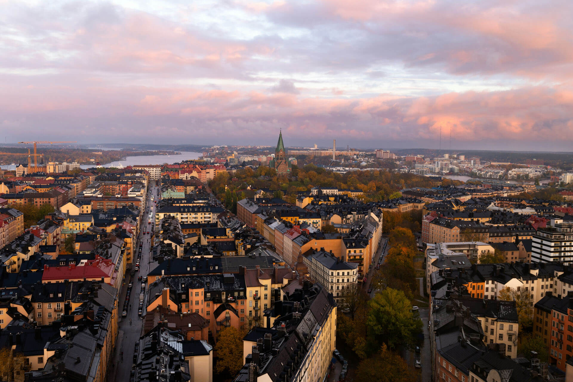 Drönarbild över tät stadsmiljö med färgglada byggnader och vatten i bakgrunden, fotograferad i varmt kvällsljus med mjuka moln över himlen.