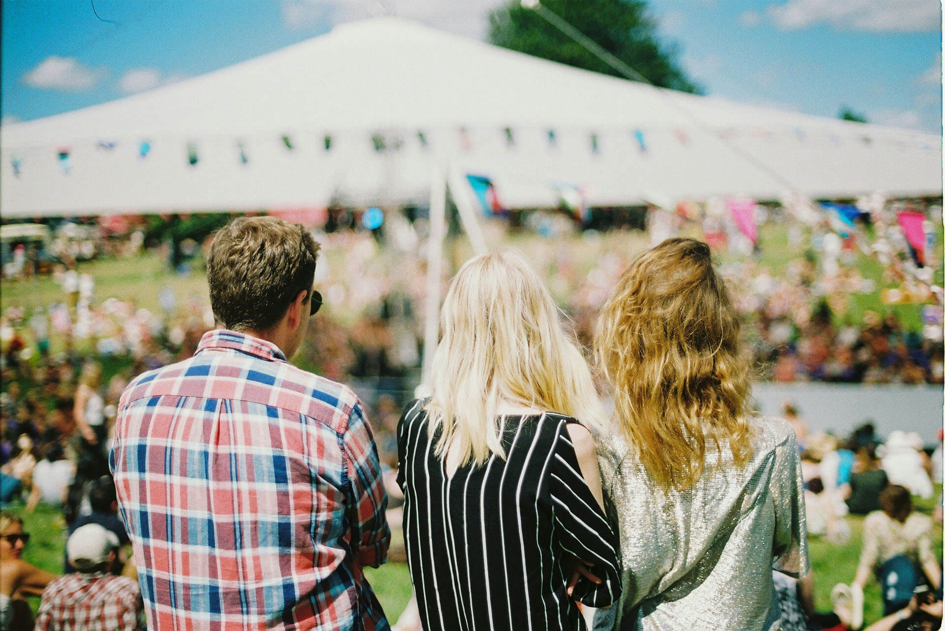 Tre personer står med ryggen mot kameran och blickar ut över stor publik framför en scen i dagsljus på en festival.