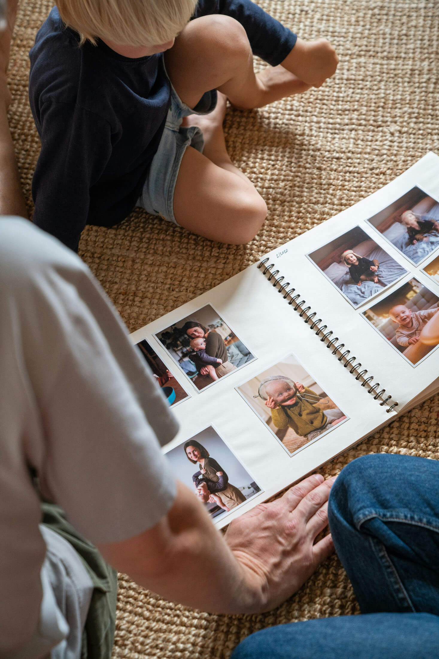  Barn sitter på en matta och bläddrar tillsammans med en vuxen i ett fotoalbum fyllt med familjebilder, fotograferat i mjukt naturligt ljus.