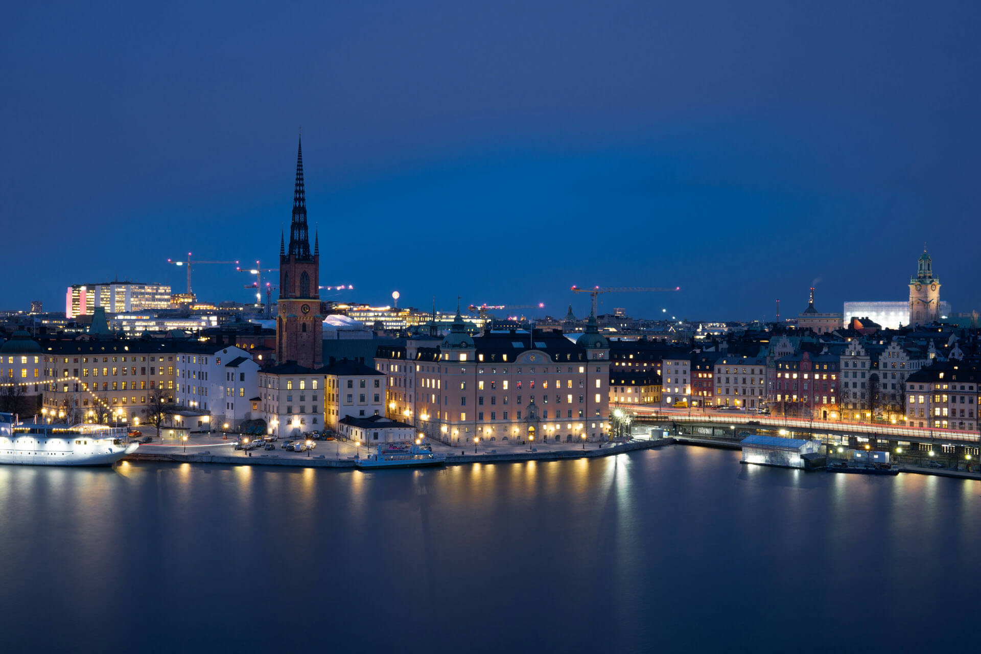 Kvällsbild över stockholm vid vatten med upplysta byggnader och kyrktorn, fotograferad i blå timmen med reflektioner i vattnet.
