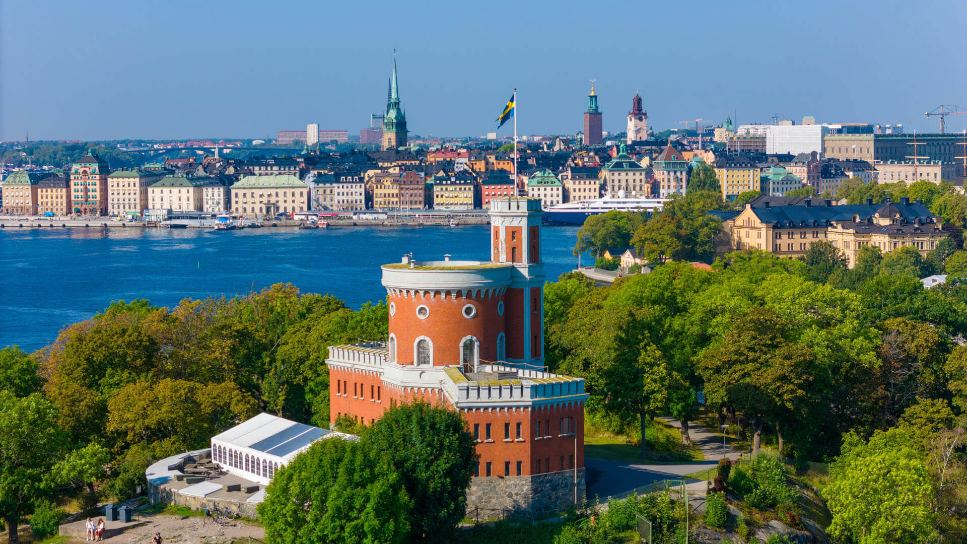 Utsikt över stad och vatten med historiska byggnader i förgrunden och ett torn med flagga omgivet av grönska under klart dagsljus.