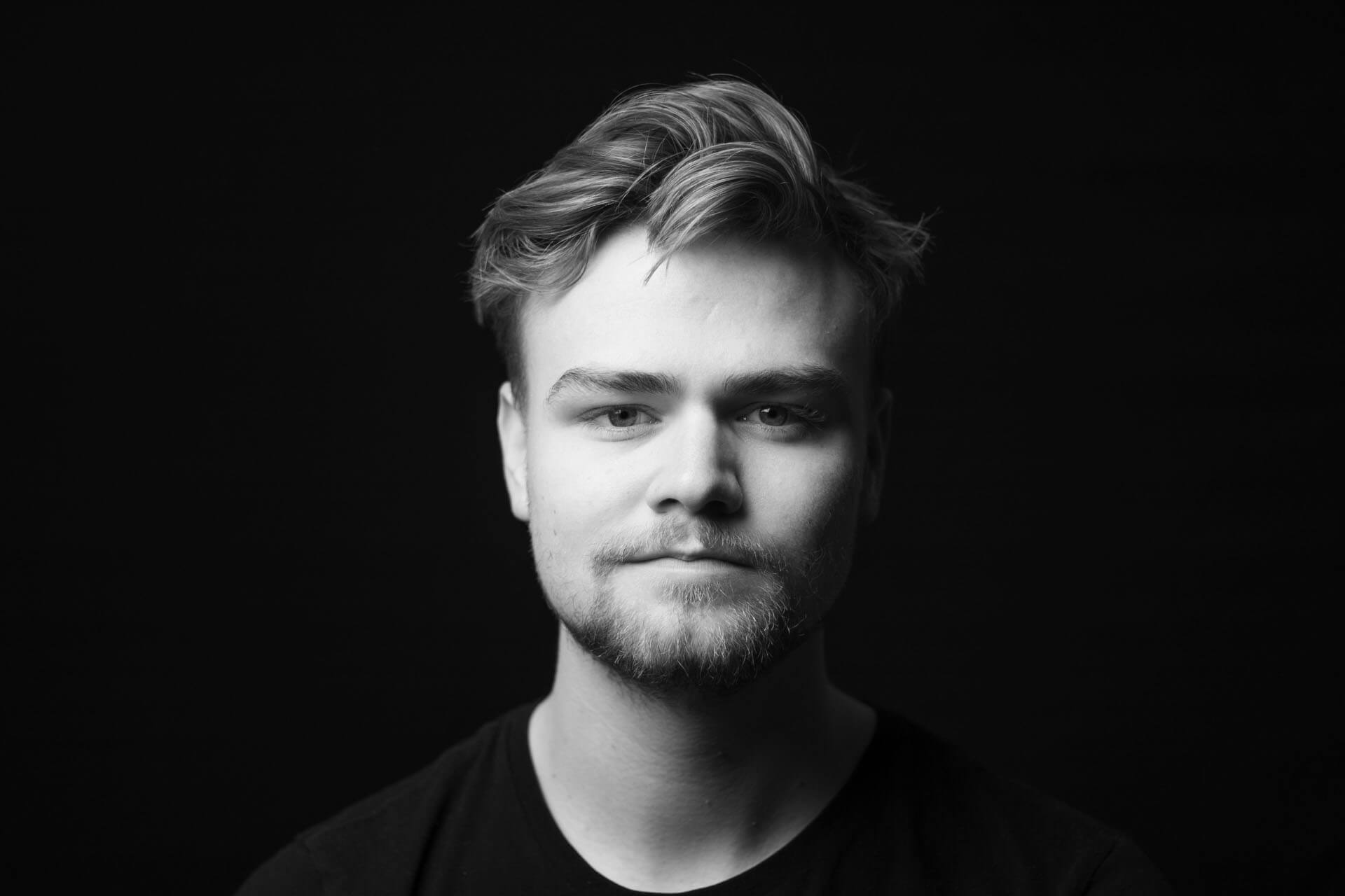Studioporträtt i svartvitt med mörk bakgrund och mjukt riktat ljus som framhäver ansikte och detaljer i en avskalad stil.