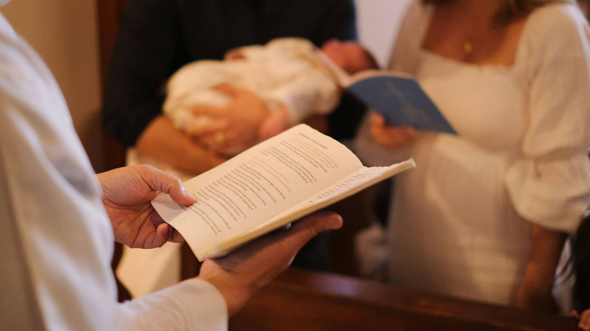 Präst håller i en bok under dopceremoni medan familj står i bakgrunden.