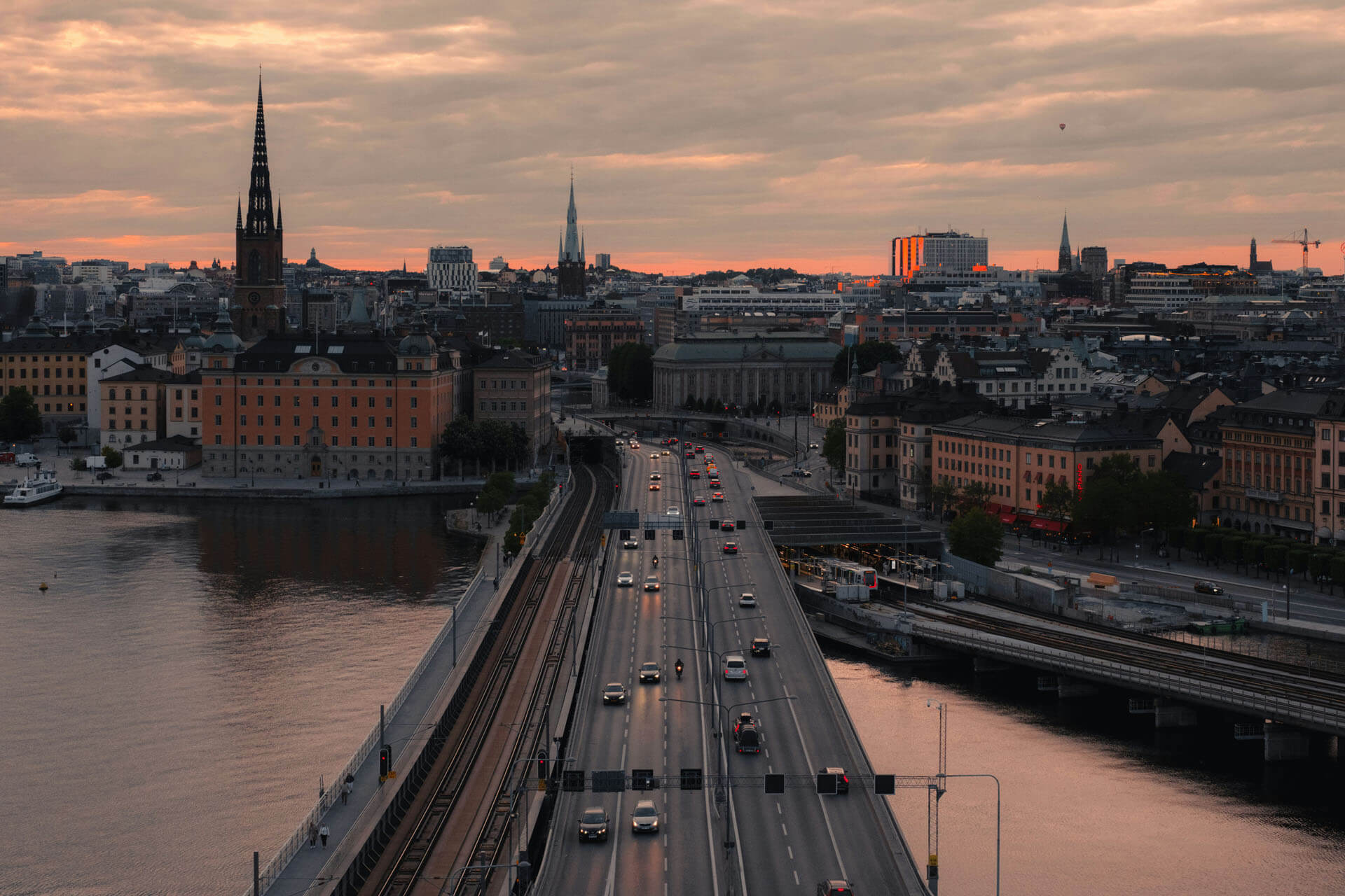 Vy över stad i solnedgång där en bred bro med trafik leder in mot centrala byggnader och kyrktorn, speglade i vattnet på båda sidor.