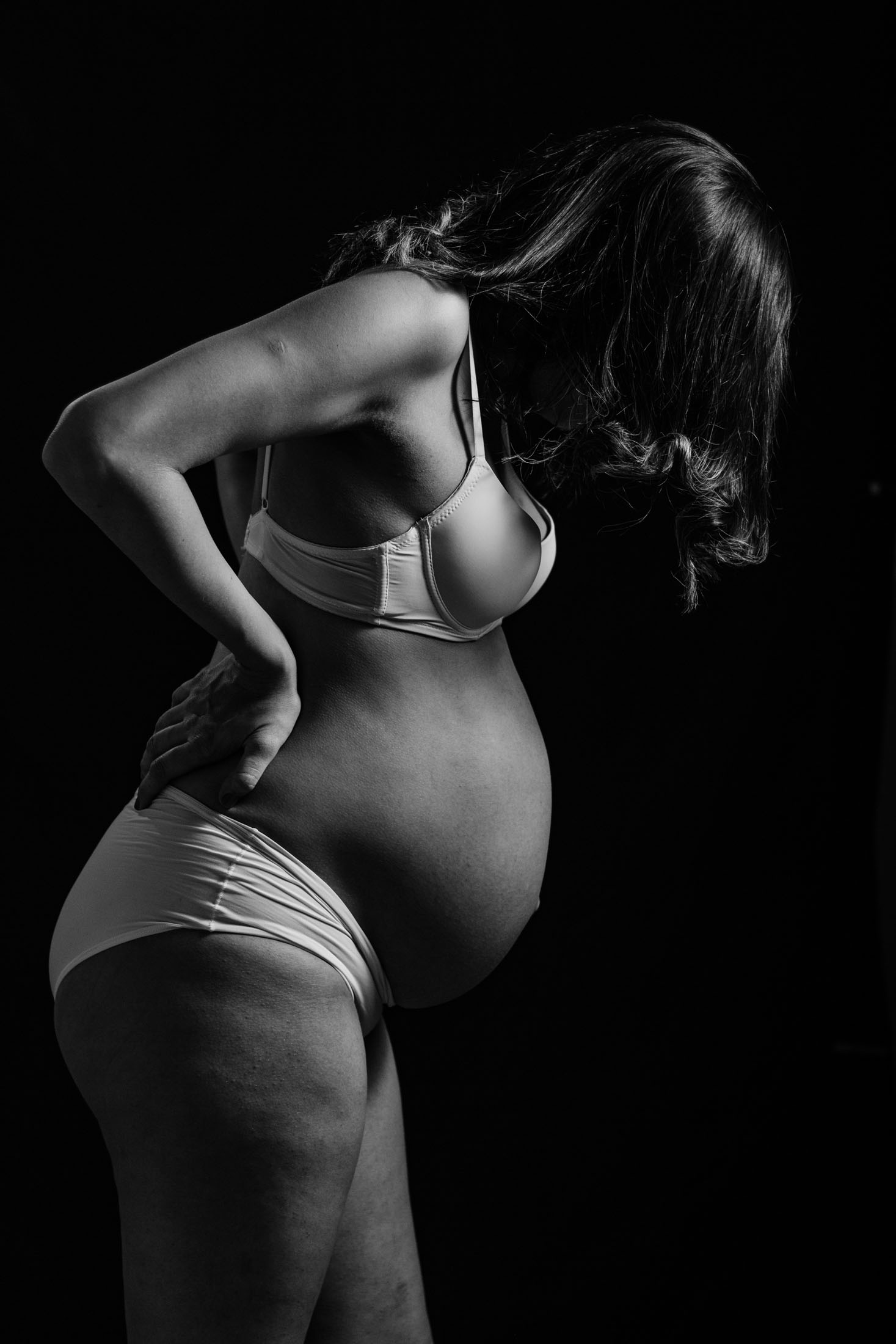 Svartvitt gravidfoto taget av fotograf i Stockholm där gravid kvinna fotograferas i sidoprofil medans hon tittar ned på magen mot mörk bakgrund. Det dramatiska ljuset skapar ett konstnärligt gravidporträtt.