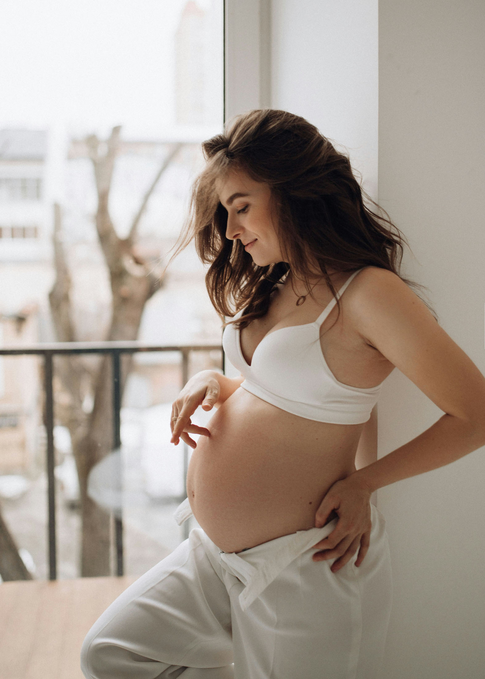 Gravidfotografering hemma i Stockholm med en gravid kvinna med mörkt hår i vit sport BH och ljusa mjukis byxor och som står vid ett öppet fönster. Det naturliga ljuset skapar en avslappnad och personlig gravidbild.