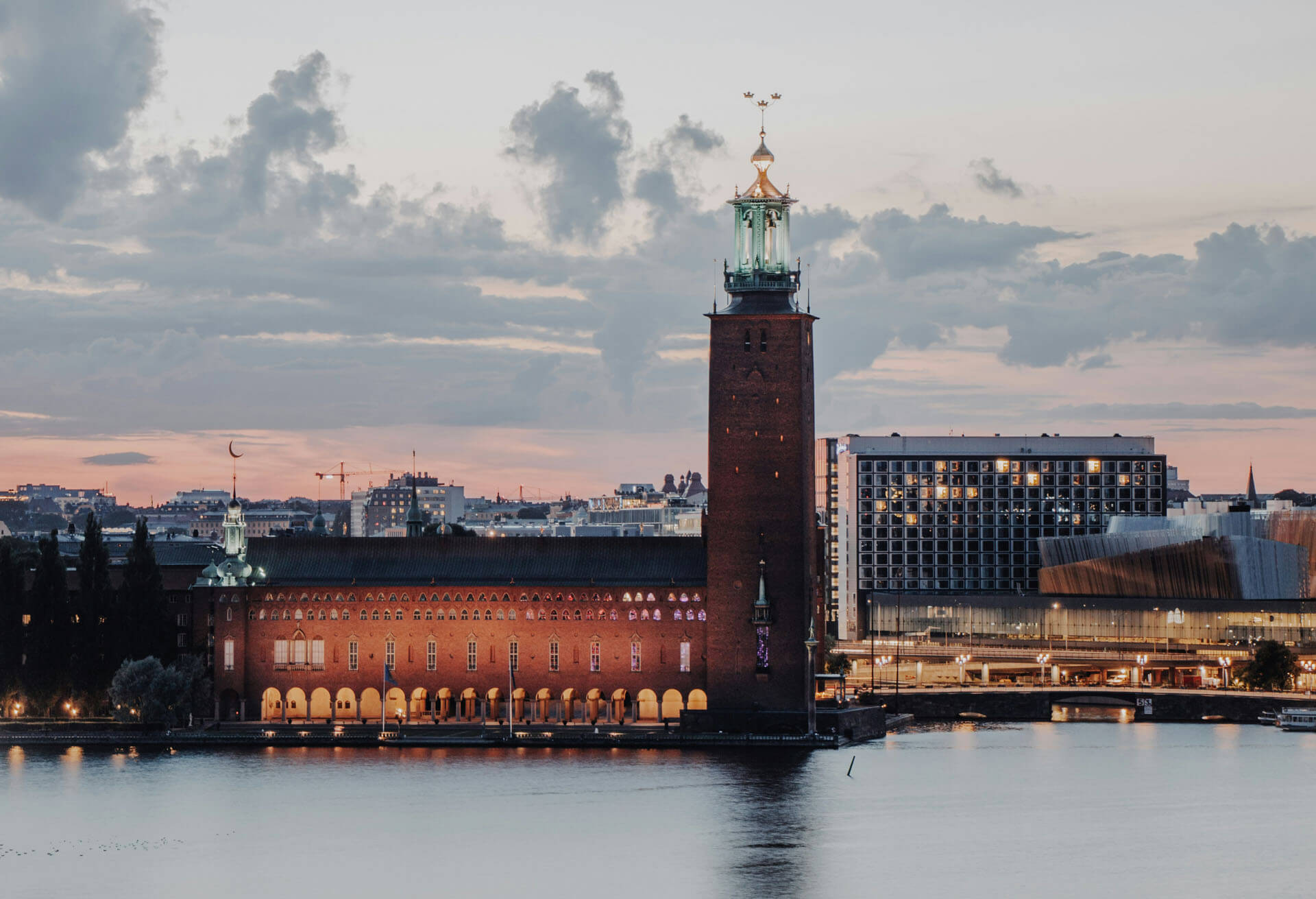 Stockholms stadshus sett över vattnet i skymningsljus. Den karakteristiska tornbyggnaden reser sig över den upplysta byggnaden längs strandkanten, medan stadens silhuett syns i bakgrunden. Det lugna vattnet speglar ljuset från byggnaderna och himlen.