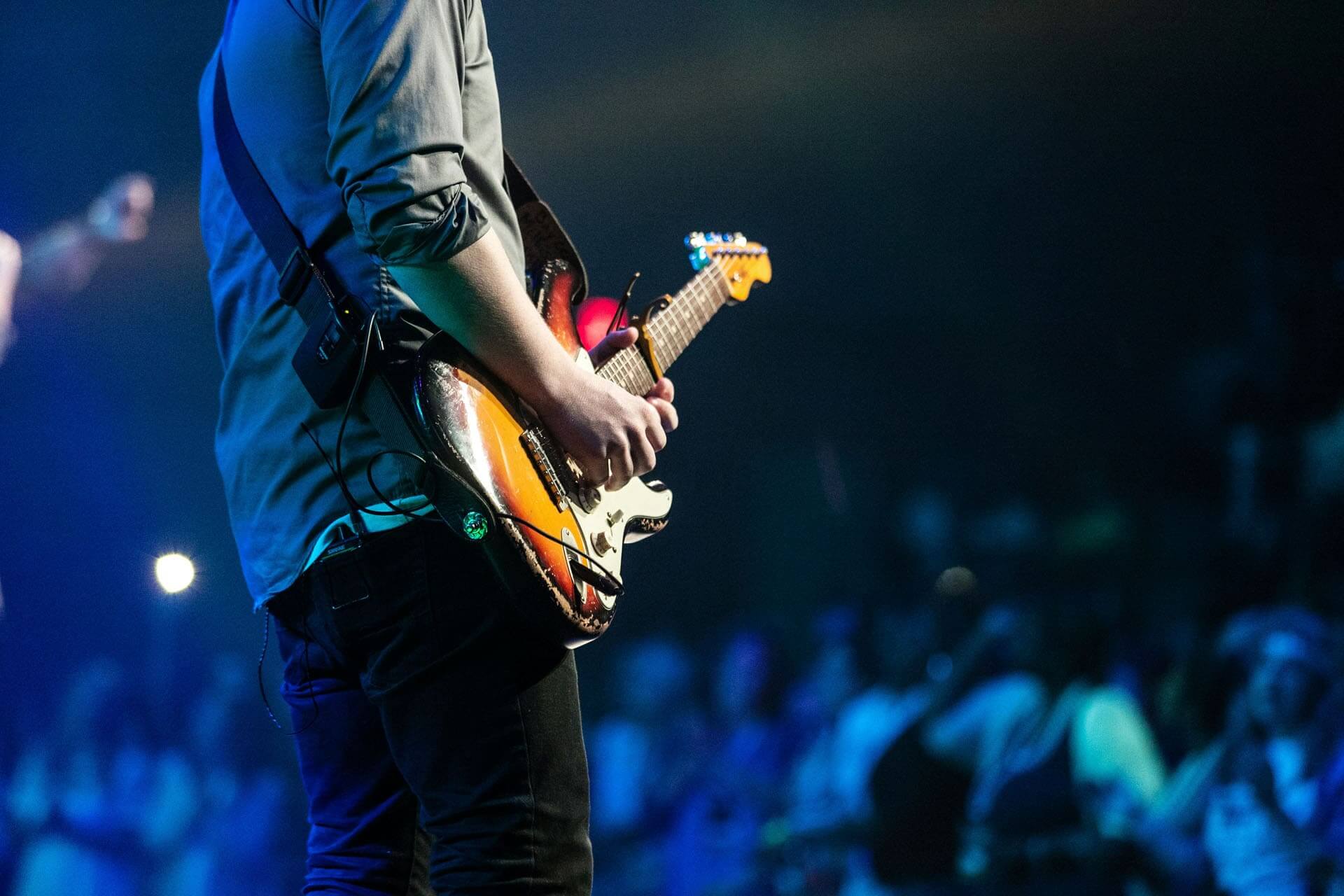 Närbild av en person som spelar elgitarr på scen, belyst av blått scenljus med publik suddigt synlig i bakgrunden.