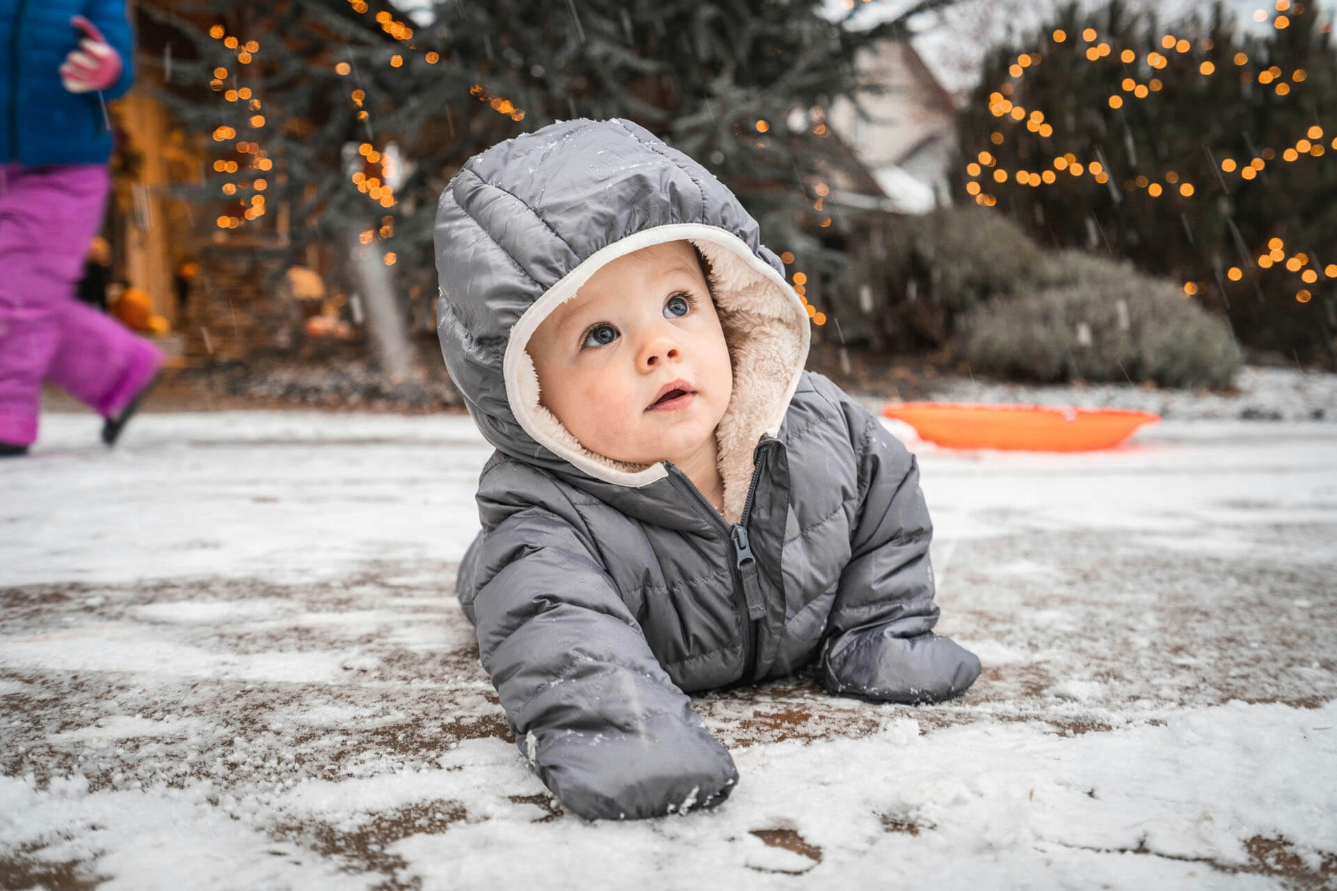 Ett spädbarn ligger på mage i snön, klätt i en grå vinteroverall och en stickad mössa med öron. Barnet tittar upp mot kameran med ett nyfiket uttryck. I bakgrunden syns vinterklädda personer och varma ljusslingor som skapar en stämningsfull utomhusmiljö.