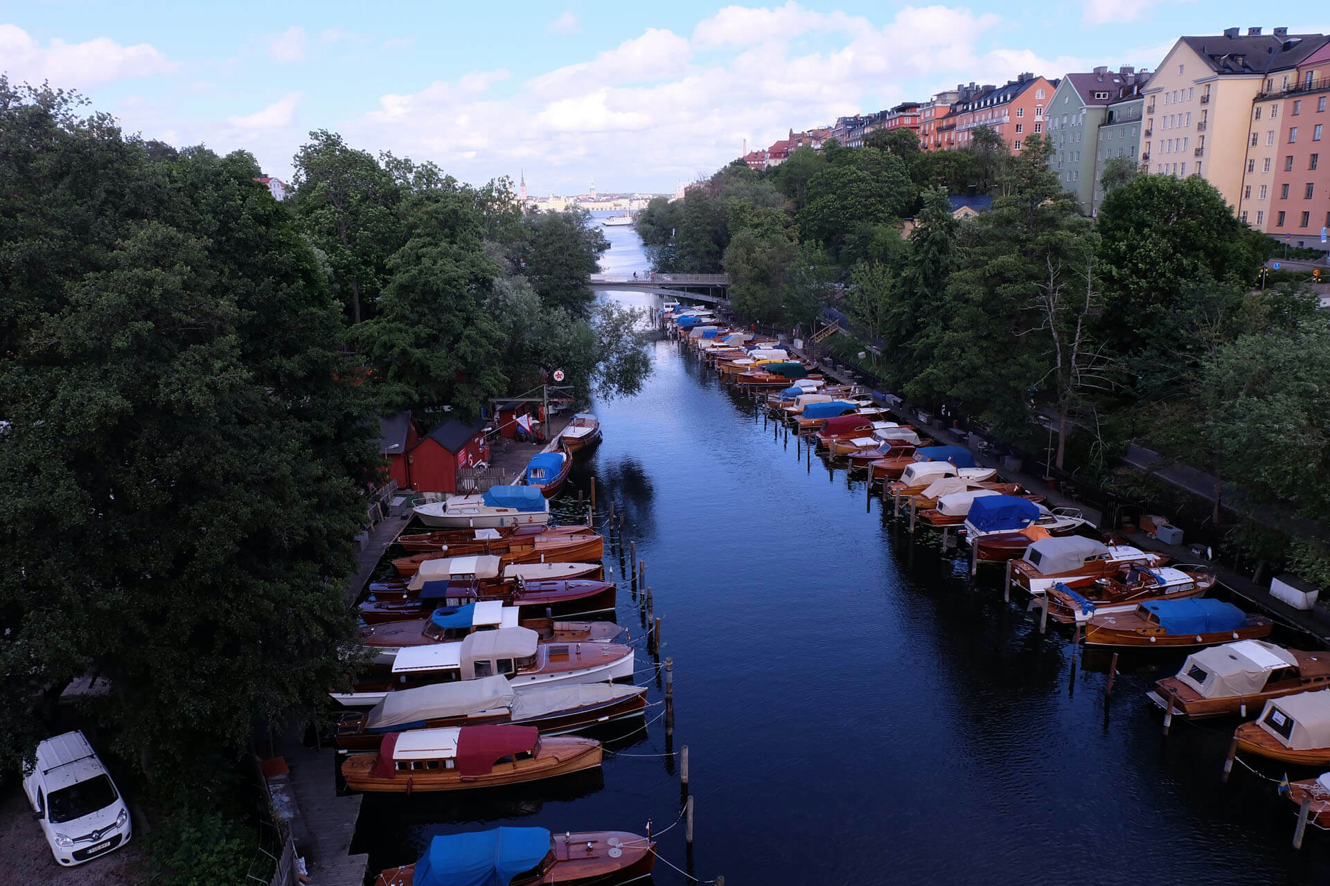 Kanal kantad av träd och färgglada byggnader med små båtar förtöjda längs båda sidor, fotograferad i mjukt kvällsljus.