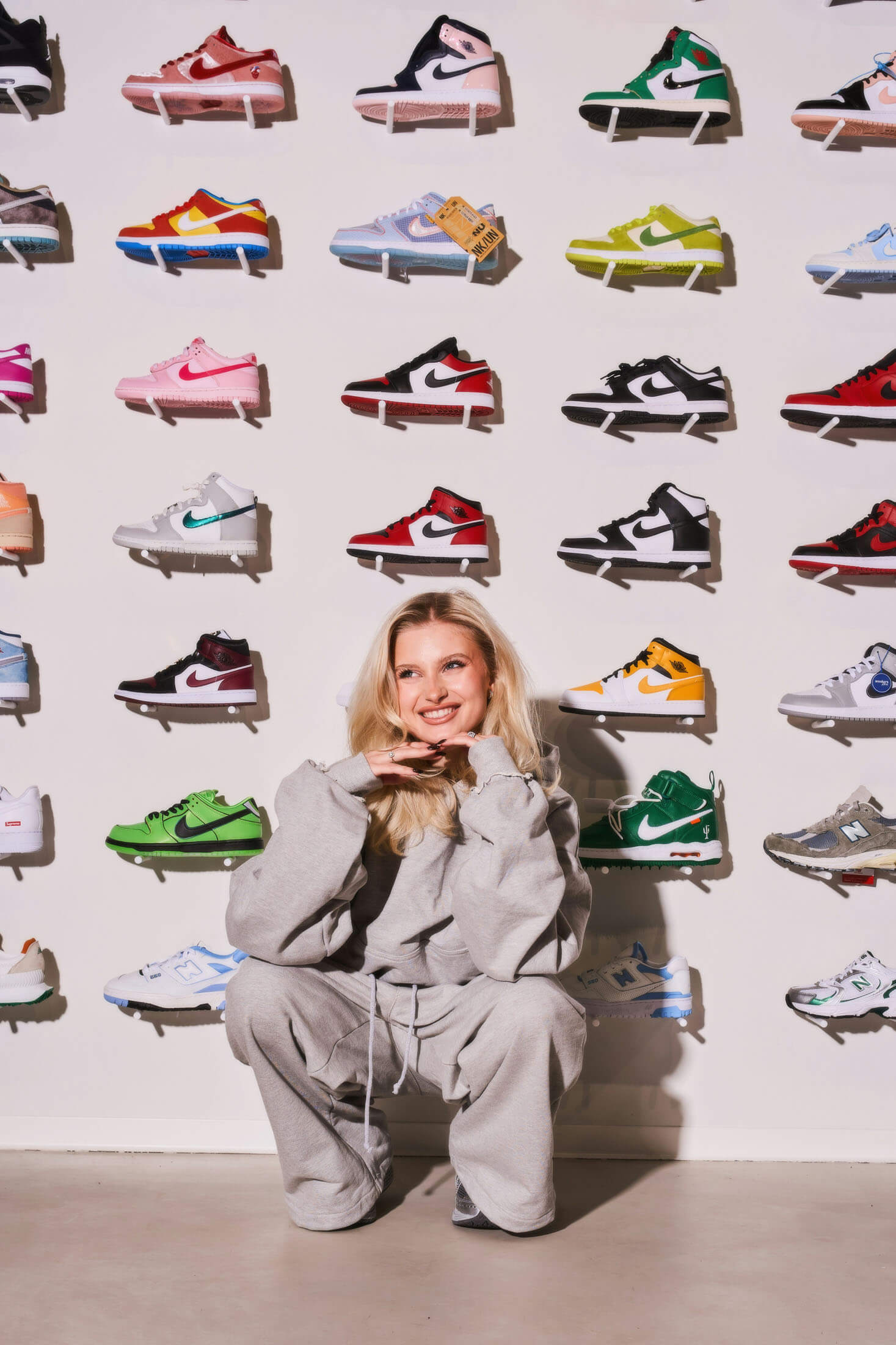 Fotografering i Stockholm där person sitter avslappnat på golvet framför en vägg med färgglada sneakers. Personen lutar sig lätt framåt med armarna vilande på knäna och blickar mot kameran. Den framåtlutad posen, den direkta blicken och den grafiska bakgrunden skapar ett modernt kampanjfoto för mode- och livsstilsvarumärken.