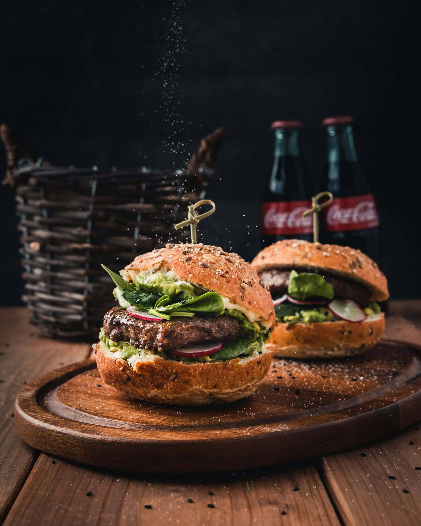 Fotografering i Stockholm där två hamburgare ligger på en träbricka med dryckesflaskor från Coca Cola i bakgrunden. Kryddor strös ovanifrån och den mörka miljön skapar ett professionellt reklamfoto för restaurang och marknadsföring.