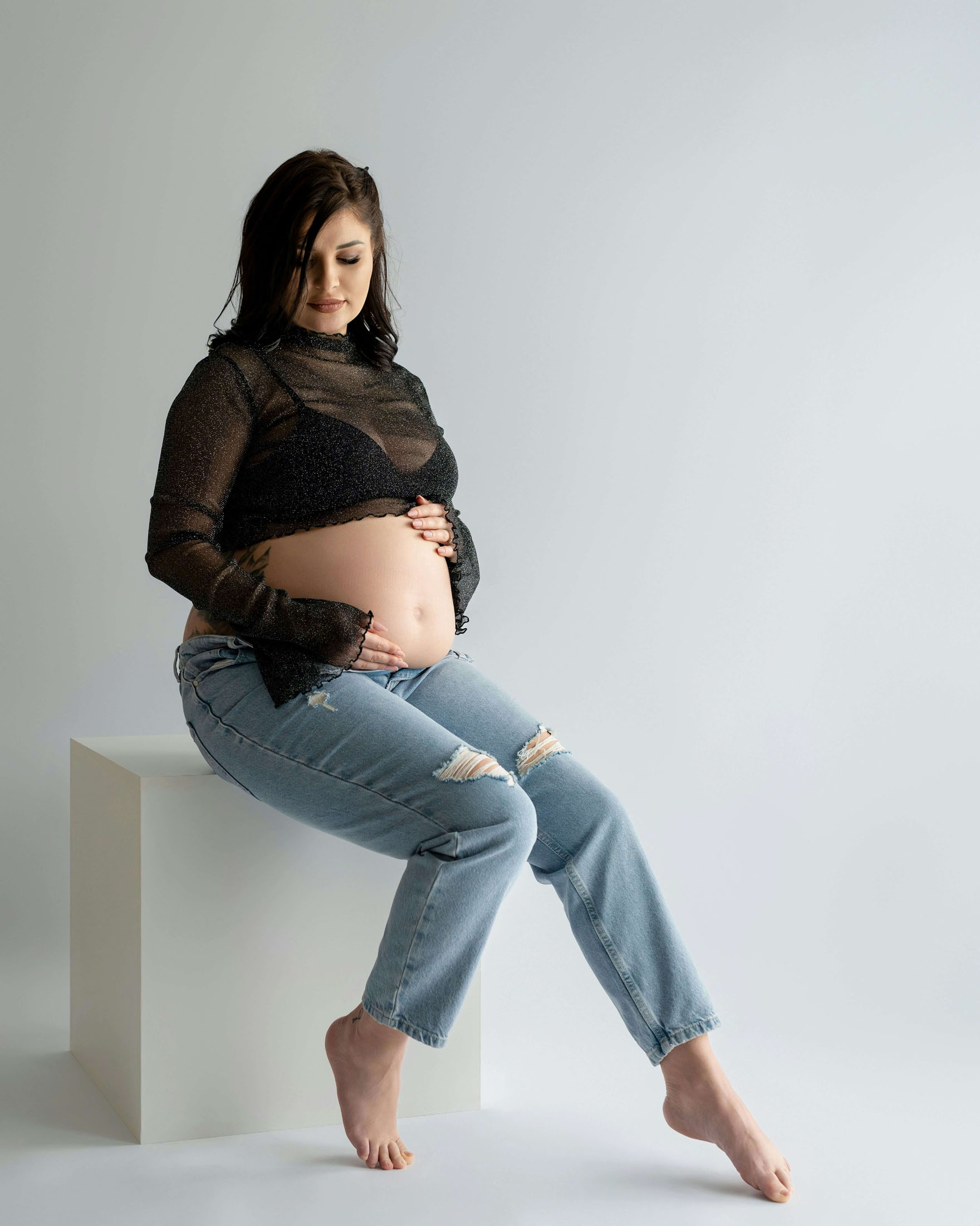 Gravidfotografering i studio i Stockholm där en gravid kvinna sitter långt fram på en vit kub med tårna mot golvet framför en ljus bakgrund. Klädd i jeans och svart topp som framhäver gravidmagen poserar hon i en avslappnad och naturlig stil.