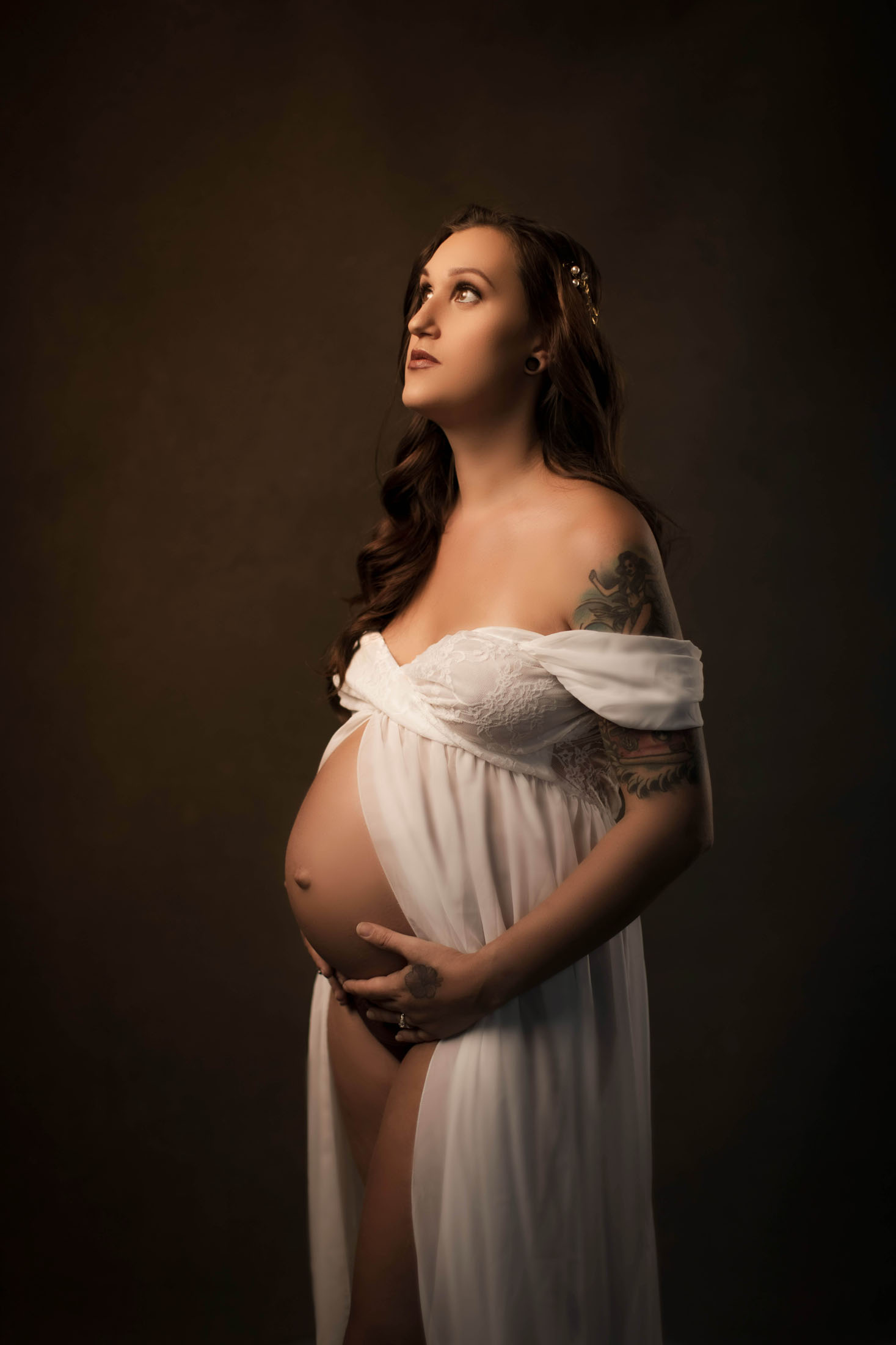 Gravidfotografering i studio där en gravid kvinna i en vit, draperad klänning som täcker de intima delarna fotograferas i profil mot en mörk bakgrund. Med händerna vilande  på en bar magen och en varm ljussättning skapas ett elegant och tidlöst gravidporträtt.