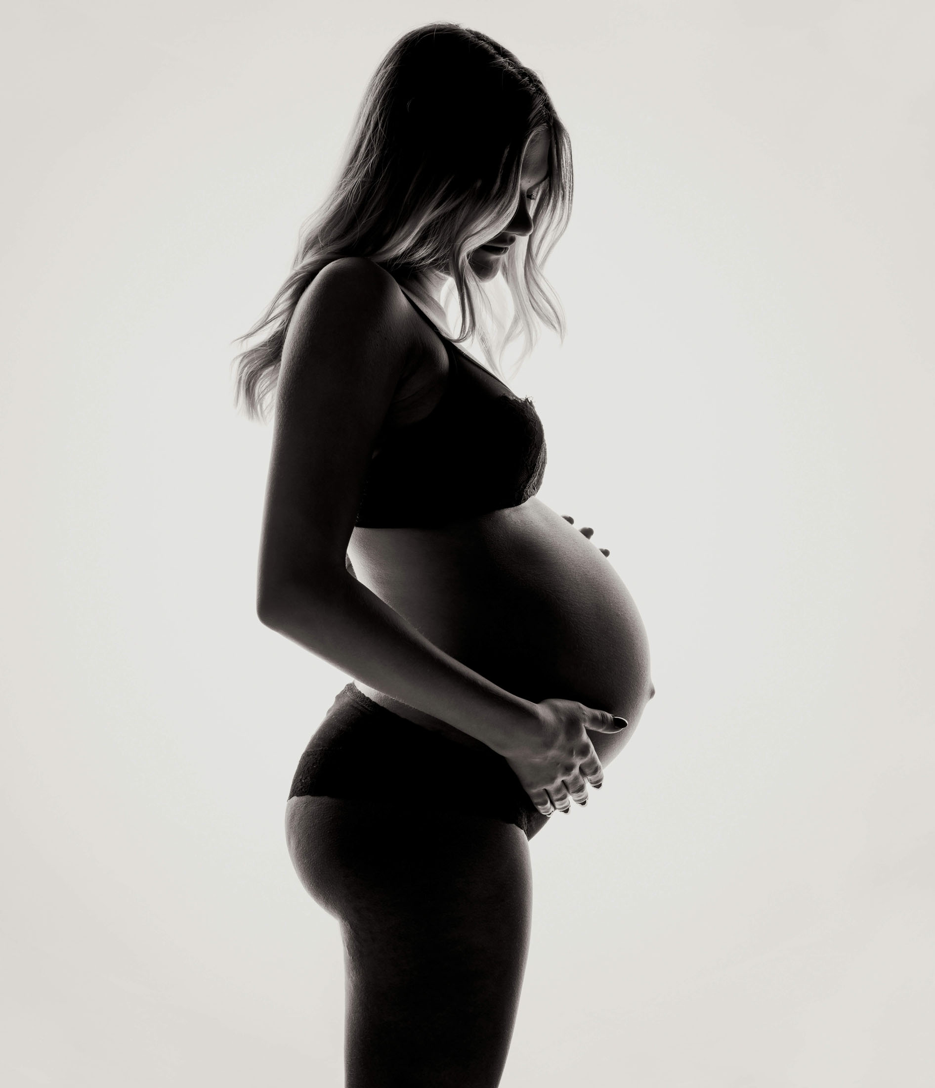  Siluettbild från gravidfotografering i studio där gravid kvinna i underkläder fotograferas mot en ljus bakgrund. Kontrasterna framhäver gravidmagens form i ett stilrent gravidfoto.