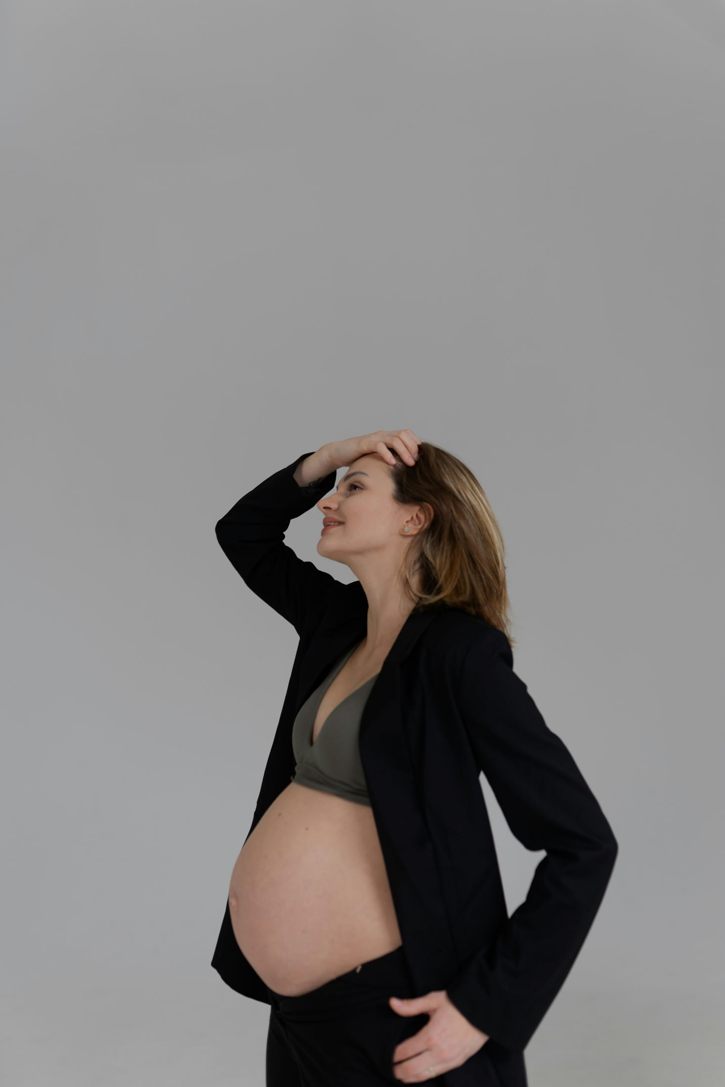 Gravidfotografering i studio i Stockholm där en gravid kvinna poserar i en öppen svart kavaj som framhäver gravidmagen. Med ena handen i håret och kroppen lätt vinklad skapas ett modernt, elegant och självsäkert gravidporträtt mot en minimalistisk bakgrund.