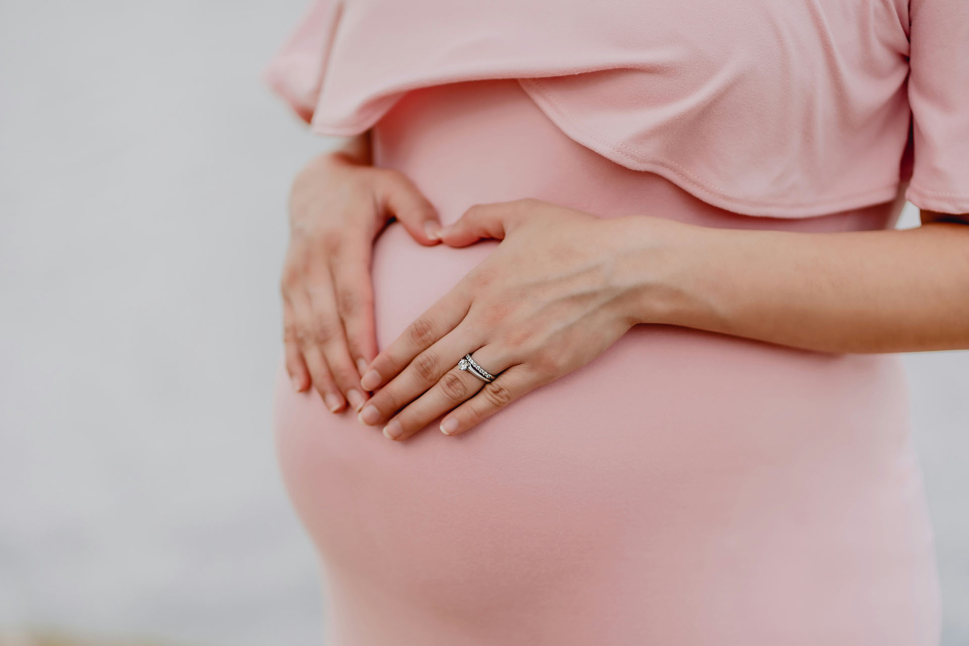 Närbild från gravidfotografering i studio där händer formar ett hjärta över gravidmagen som täcks av rosa klänning. Detaljen skapar en personlig gravidbild med fokus på väntan.
