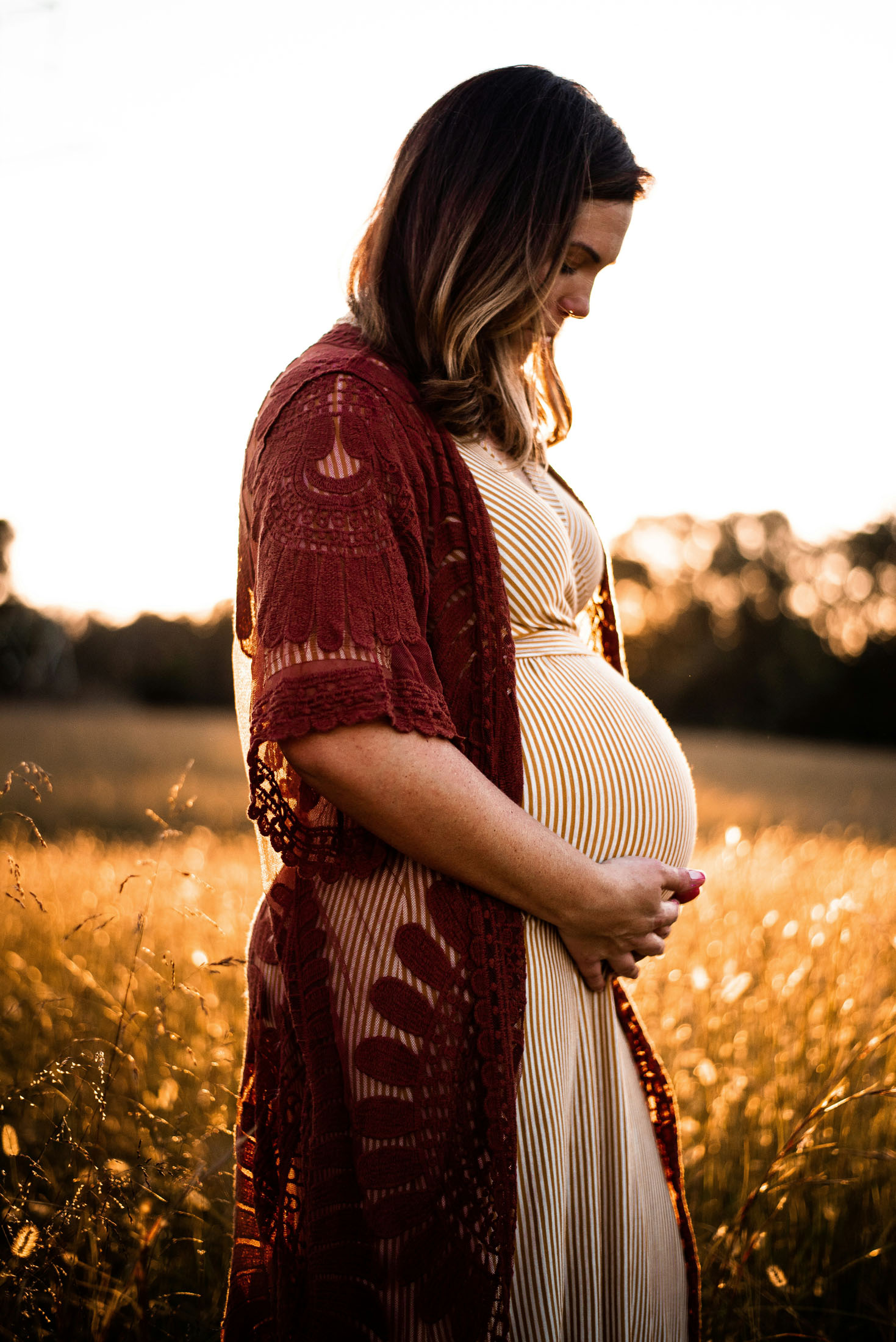 Gravidfotografering utomhus i Stockholm där en gravid kvinna står i högt gräs och blickar ner mot sin gravidmage. Klädd i en ljus klänning och röd spetskimono håller hon händerna på magen medan det gyllene kvällsljuset skapar ett varmt och stämningsfullt gravidporträtt