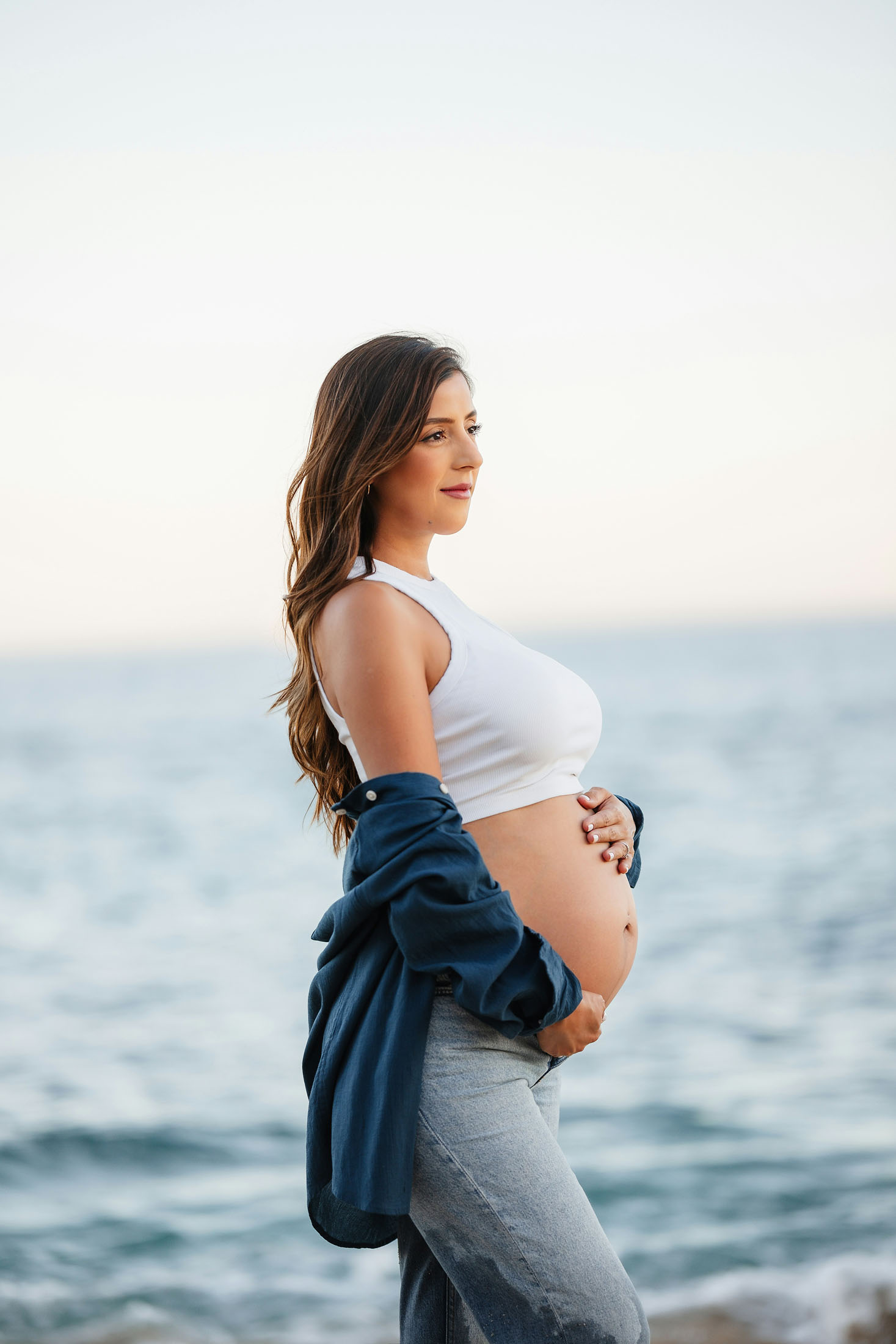 Gravidfotografering utomhus i Stockholm där en gravid kvinna står i profil vid havet och håller händerna på magen. En jeansjacka vilar över armarna medan det mjuka kvällsljuset framhäver gravidmagen i ett naturligt och stämningsfullt gravidporträtt.