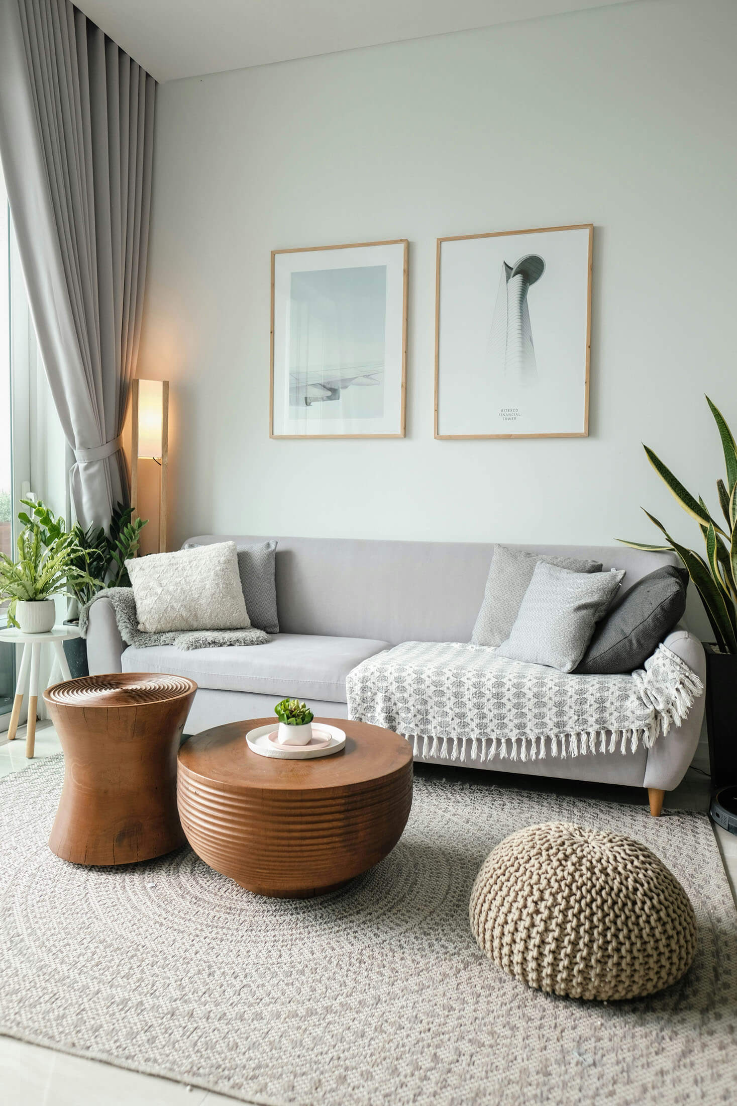Ljust vardagsrum med grå soffa, runda träbord och stora växter, där mjukt dagsljus faller in från ett fönster med gardiner och skapar en lugn, neutral färgpalett.
