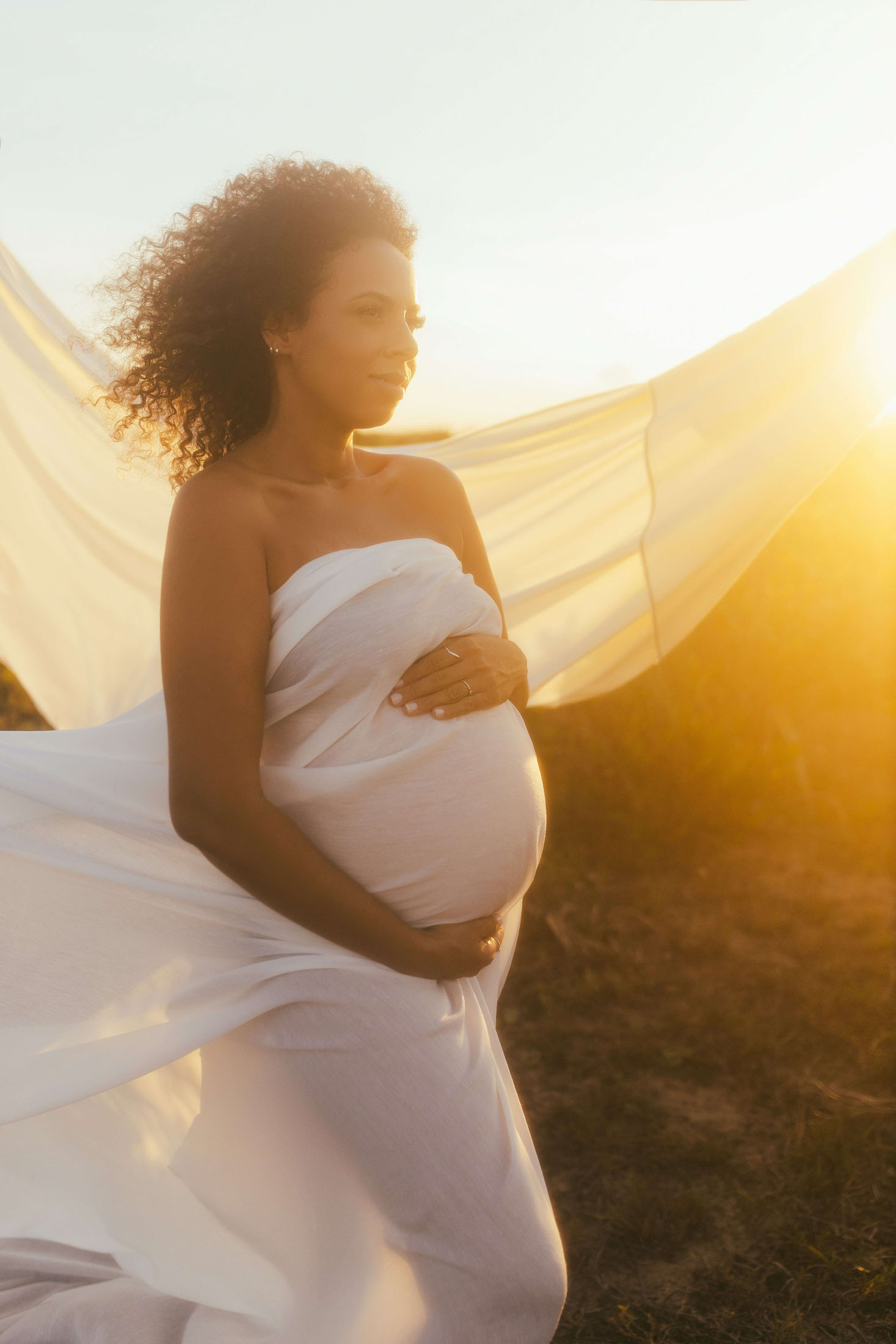 Konstnärlig gravidfotografering utomhus i Stockholm där en gravid kvinna håller händerna på magen medan det vita lakandet fångas av vinden. Det gyllene motljuset från  solnedgången tillsammans med vinden framhäver gravidmagen och skapar ett varmt, drömskt och stämningsfullt gravidporträtt.