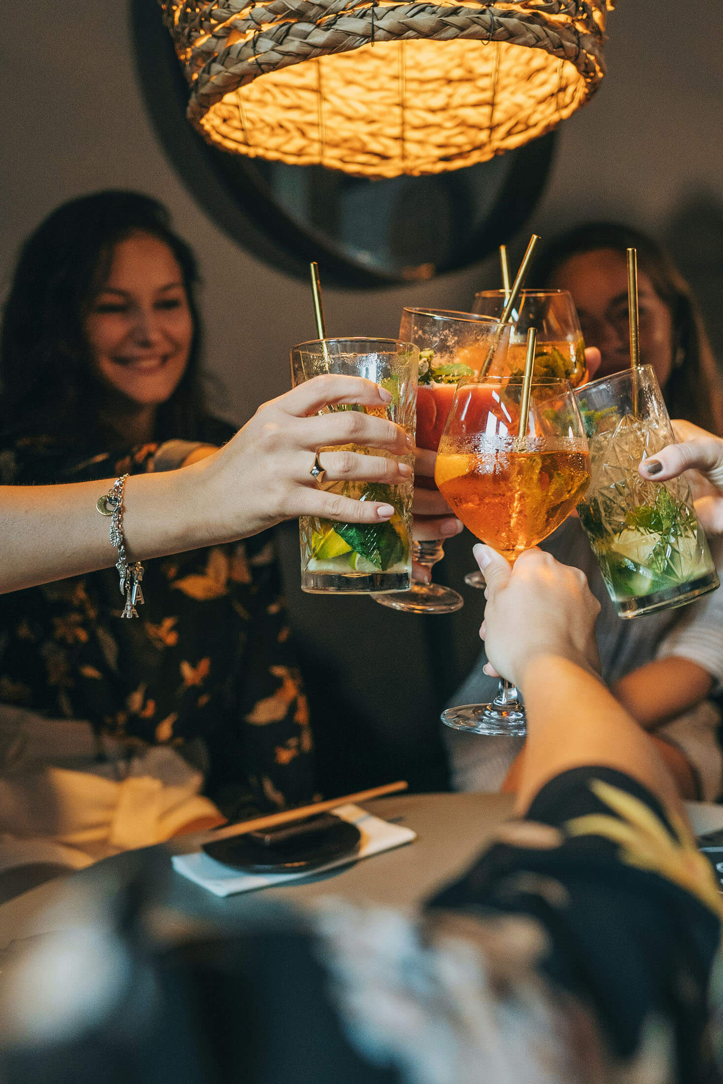 Flera personer skålar med cocktails runt ett bord i varm belysning, med lampa ovanför och mjuk bakgrundsoskärpa.