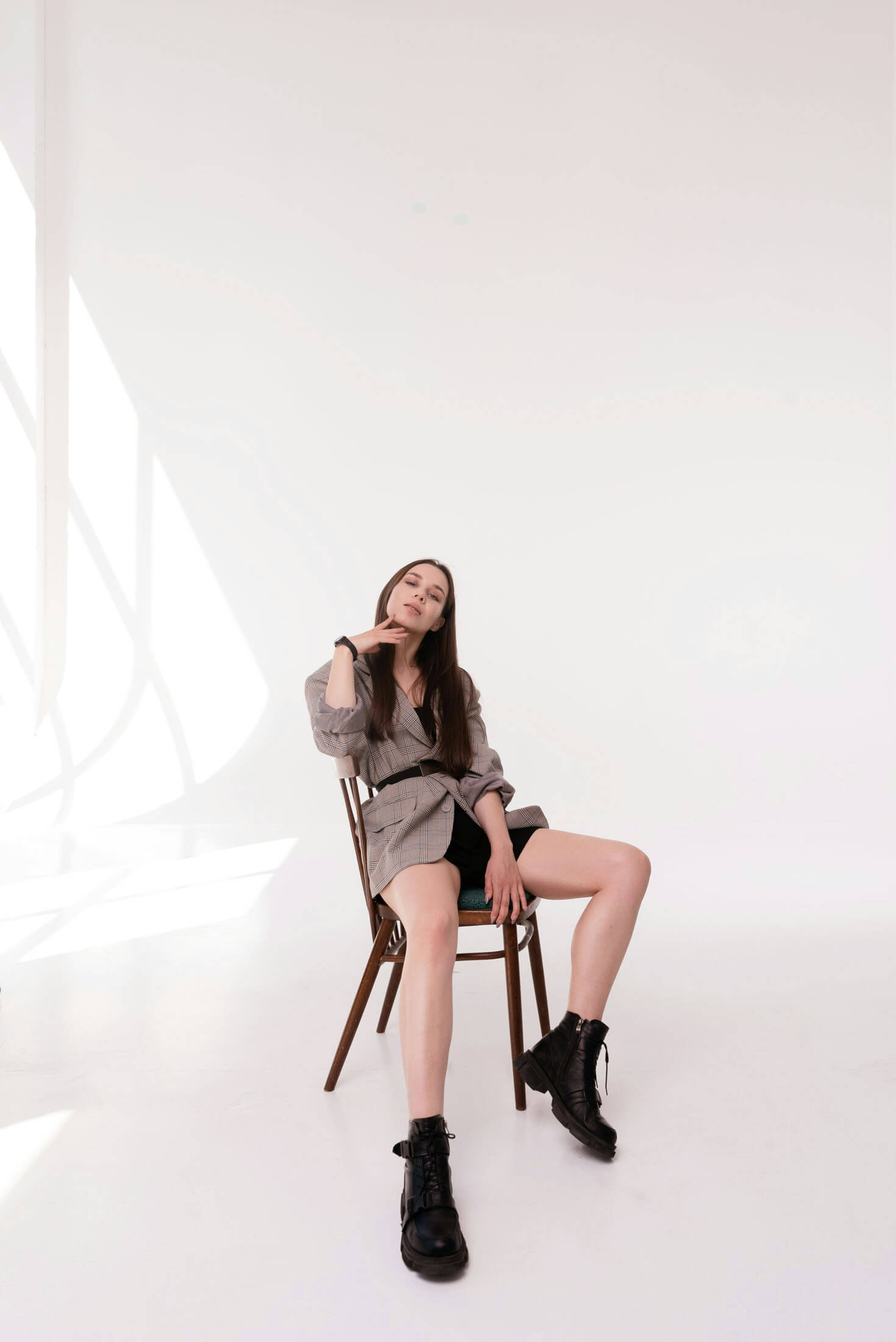 Helkroppsporträtt i studio där person sitter avslappnat på stol mot vit bakgrund, fotograferad i mjukt naturligt ljus med minimalistisk komposition.