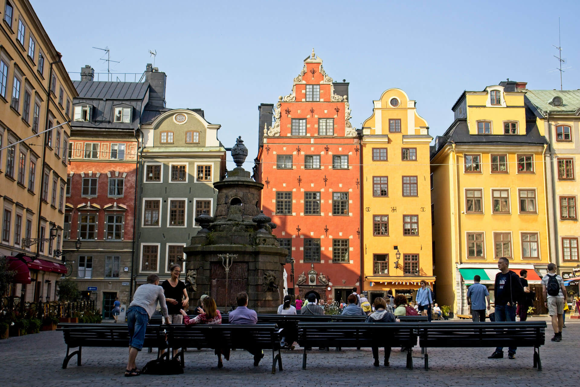 Torg i Gamla stan i Stockholm med färgglada historiska byggnader och en fontän i mitten, där människor sitter och rör sig i kvällsljus.