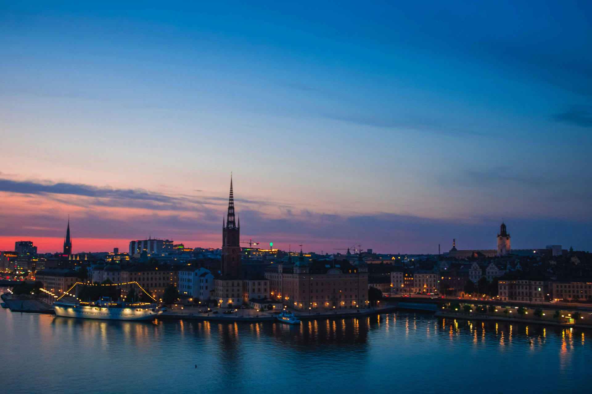 Stadsvy över Stockholm i skymning med kyrktorn, upplysta byggnader och reflektioner i vattnet under blå timmen.