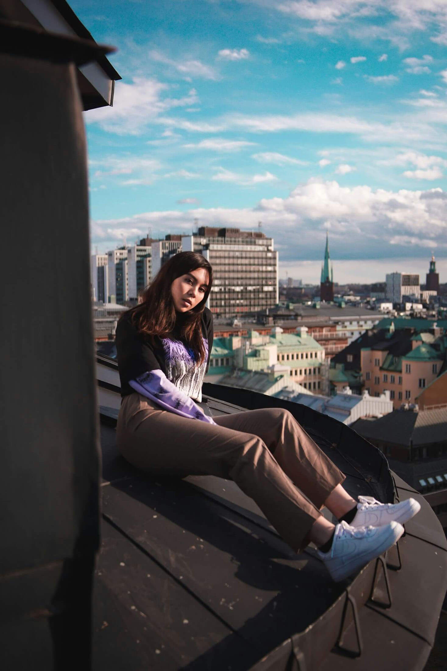 Person sitter på ett tak med utsikt över Stockholm, fotograferad i naturligt ljus med stadens byggnader och kyrktorn i bakgrunden.