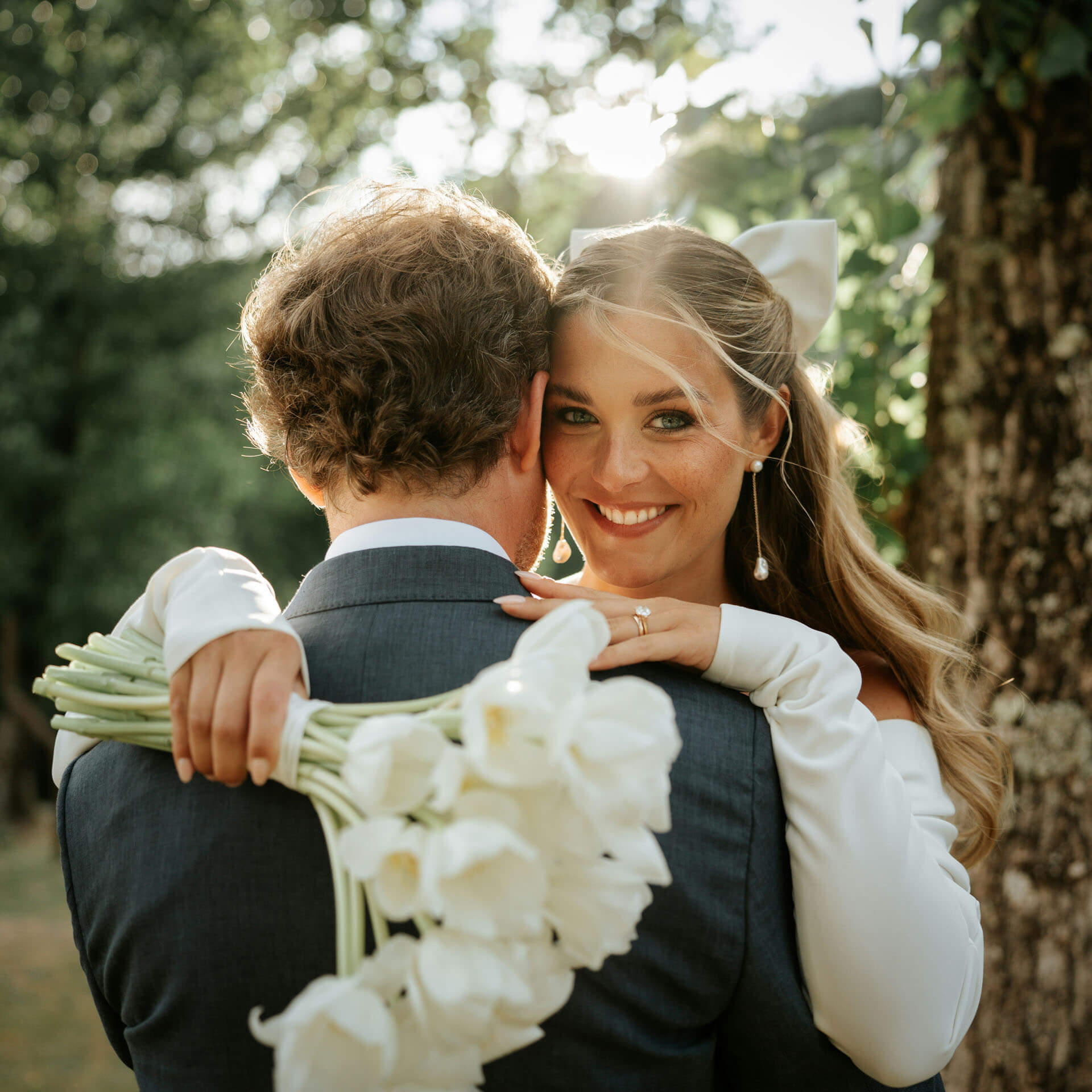 Brud håller vita blommor och omfamnar sin man i solbelyst porträtt i naturen.