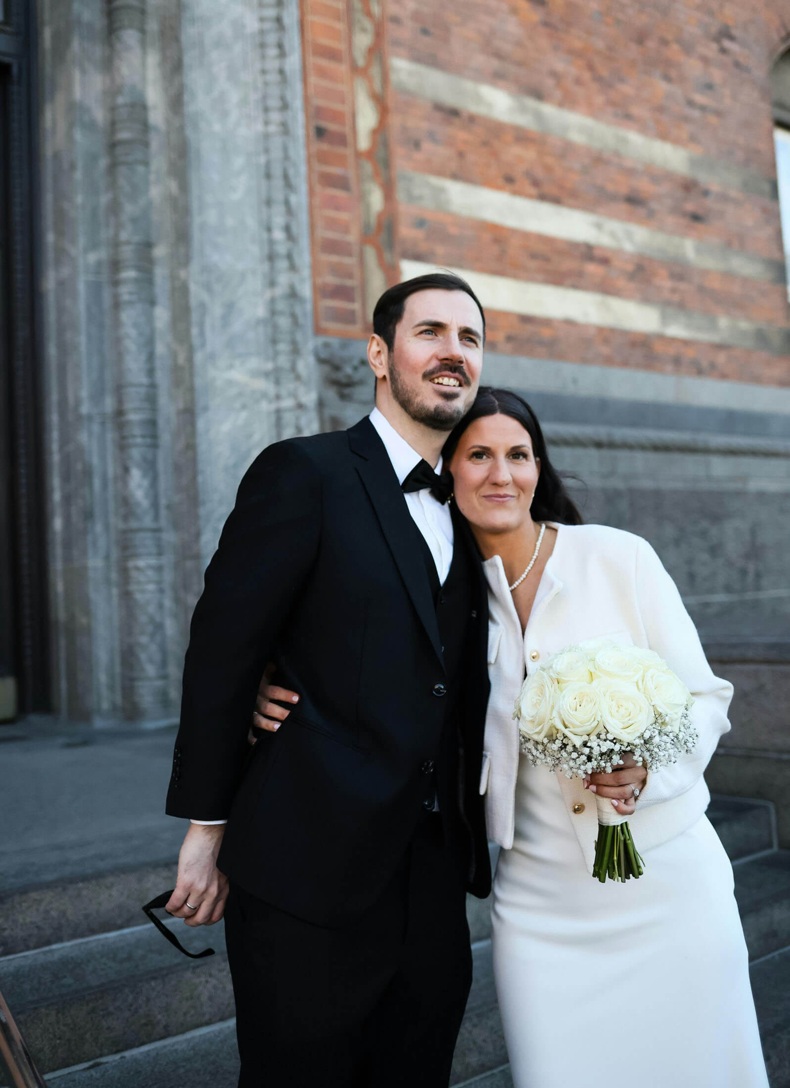 Foto på brudpar som står tätt tillsammans utanför Stockholms Stadshus var bruden håller en brudbukett medans brudgummen kramar om bruden från sidan. Tegelväggarna och det naturliga ljuset skapar ett tidlöst porträtt från en borgerlig vigsel i Stockholm, fotograferat av fotograf i Stockholm.